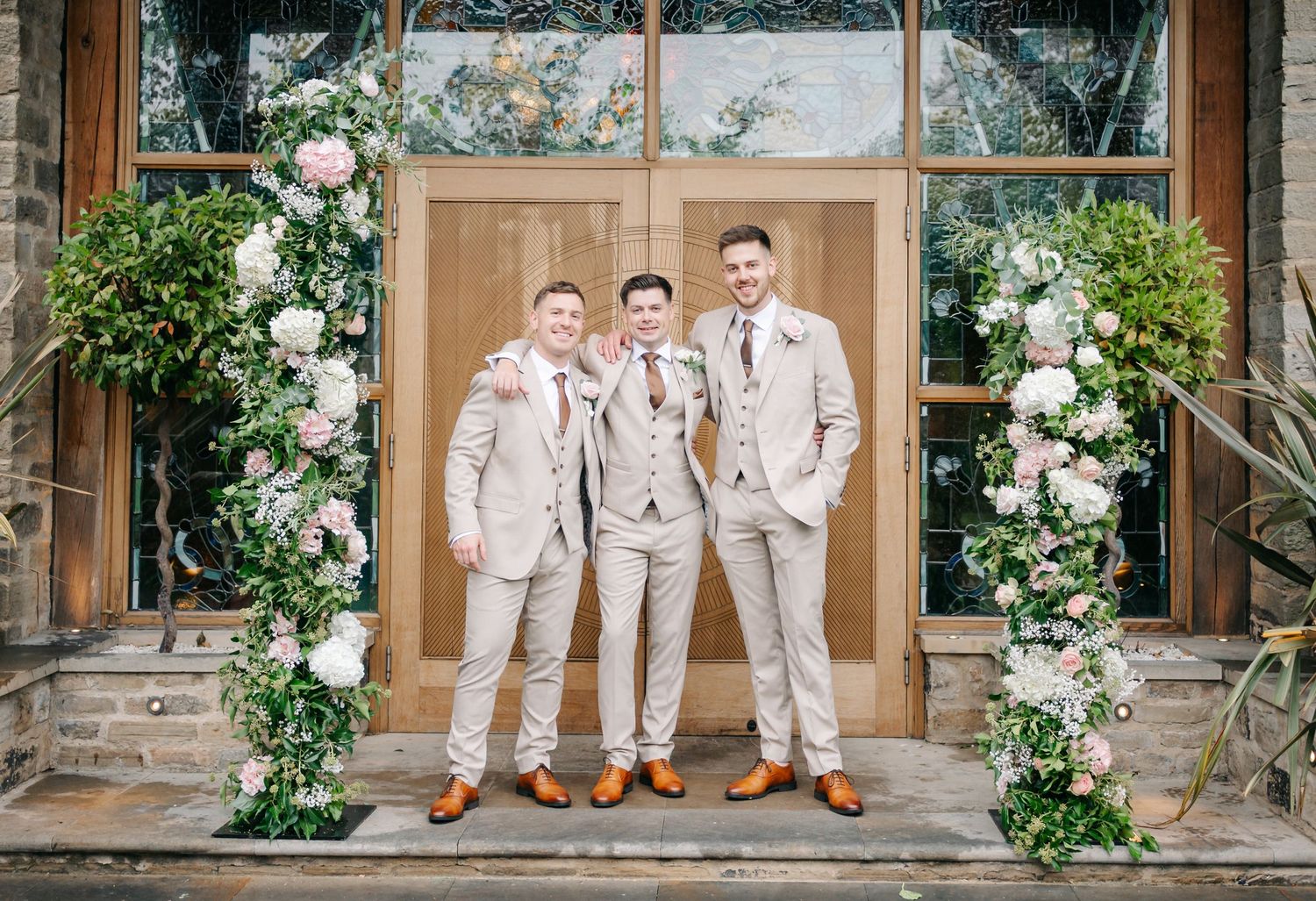 Groomsmen in beige suits and brown shoes pose together on stone steps adorned with white floral arrangements.