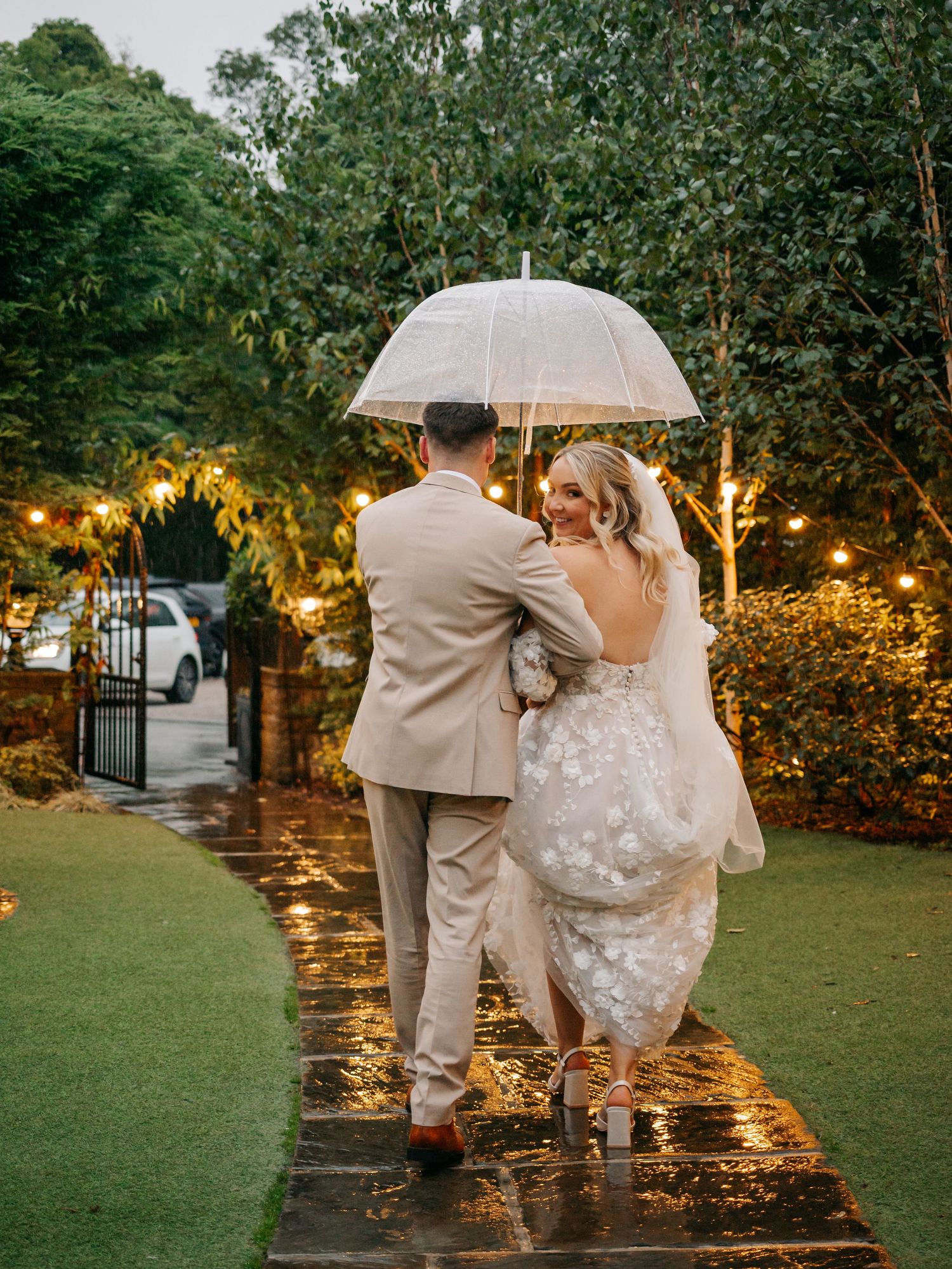 Newlyweds share romantic moment under clear umbrella on rainy garden path with twinkling lights.
