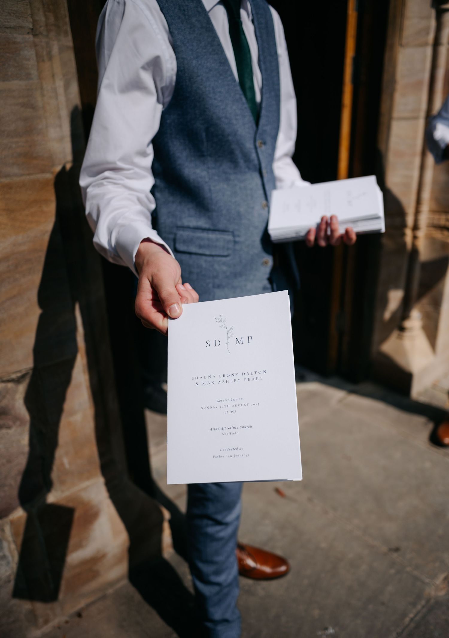 Wedding guest in grey suit holds white ceremony program while walking through a sunlit hallway.