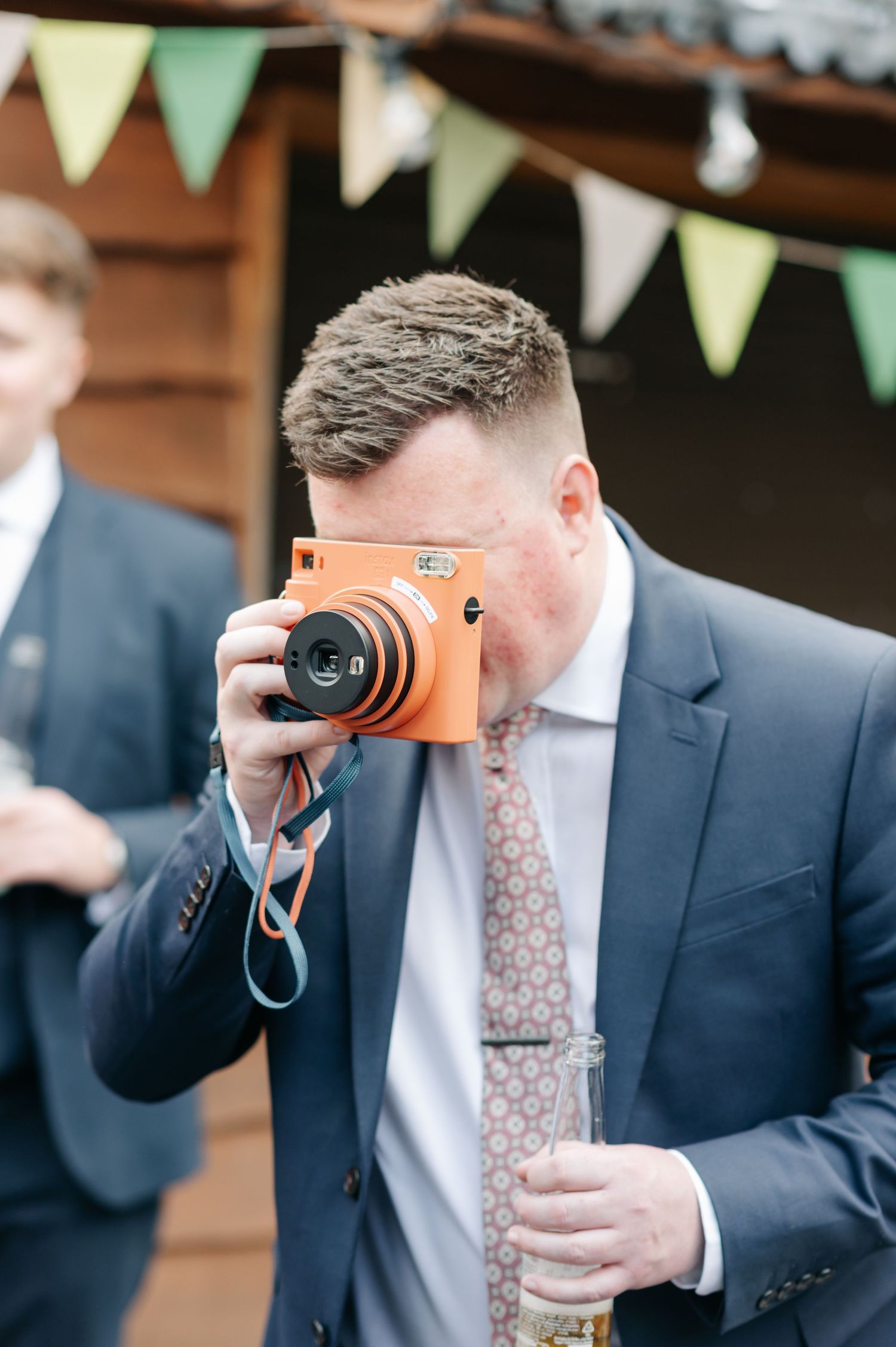 Guest in blue suit taking photo with vintage orange camera at outdoor wedding reception.