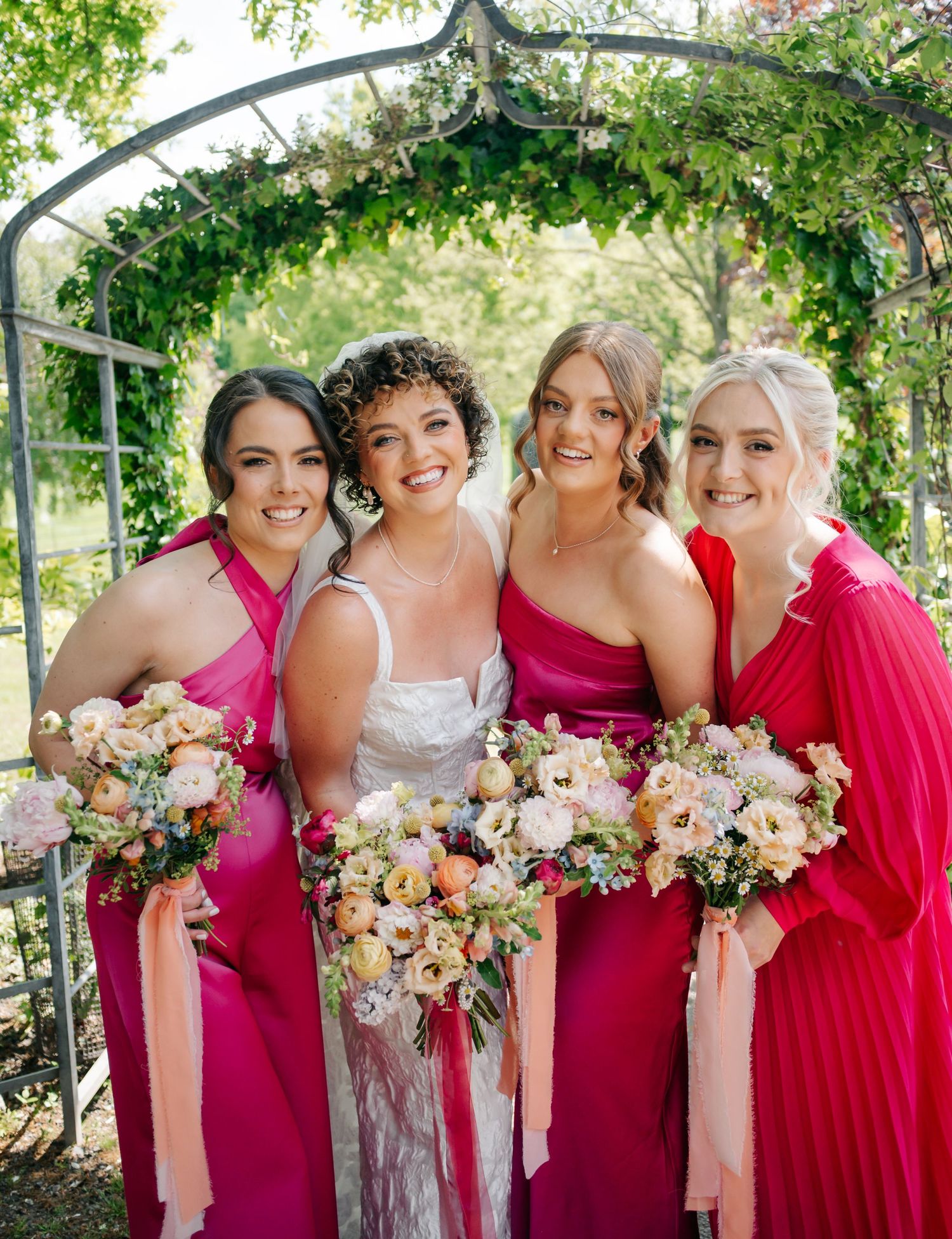 Wedding party in bright pink dresses poses together under a garden arch with floral bouquets.