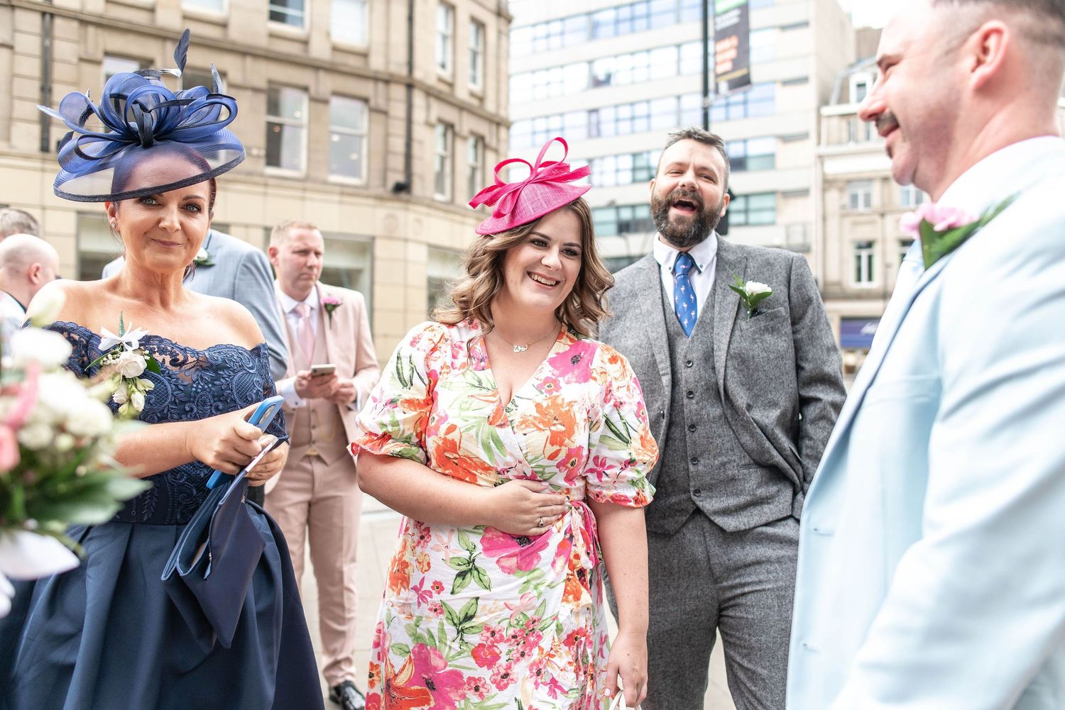 Wedding guests in colorful dresses and fascinator hats chatting outdoors.