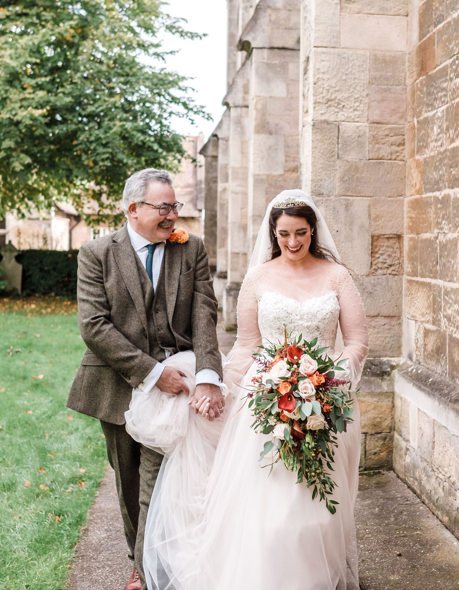 A couple walking along a stone church exterior with a beautiful bridal bouquet.