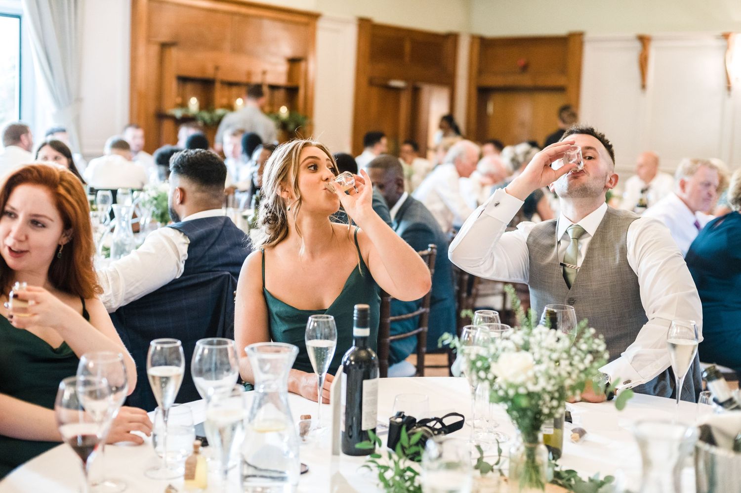 Wedding guests enjoying themselves at an elegant indoor reception with wine glasses and floral decor.