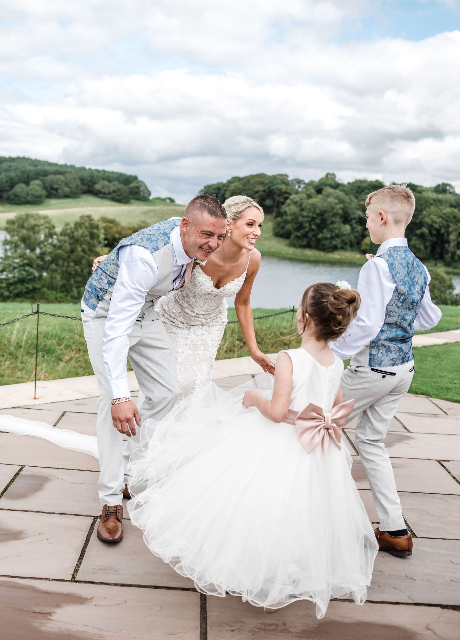 Newlyweds dancing with children on an outdoor stone patio overlooking scenic countryside.