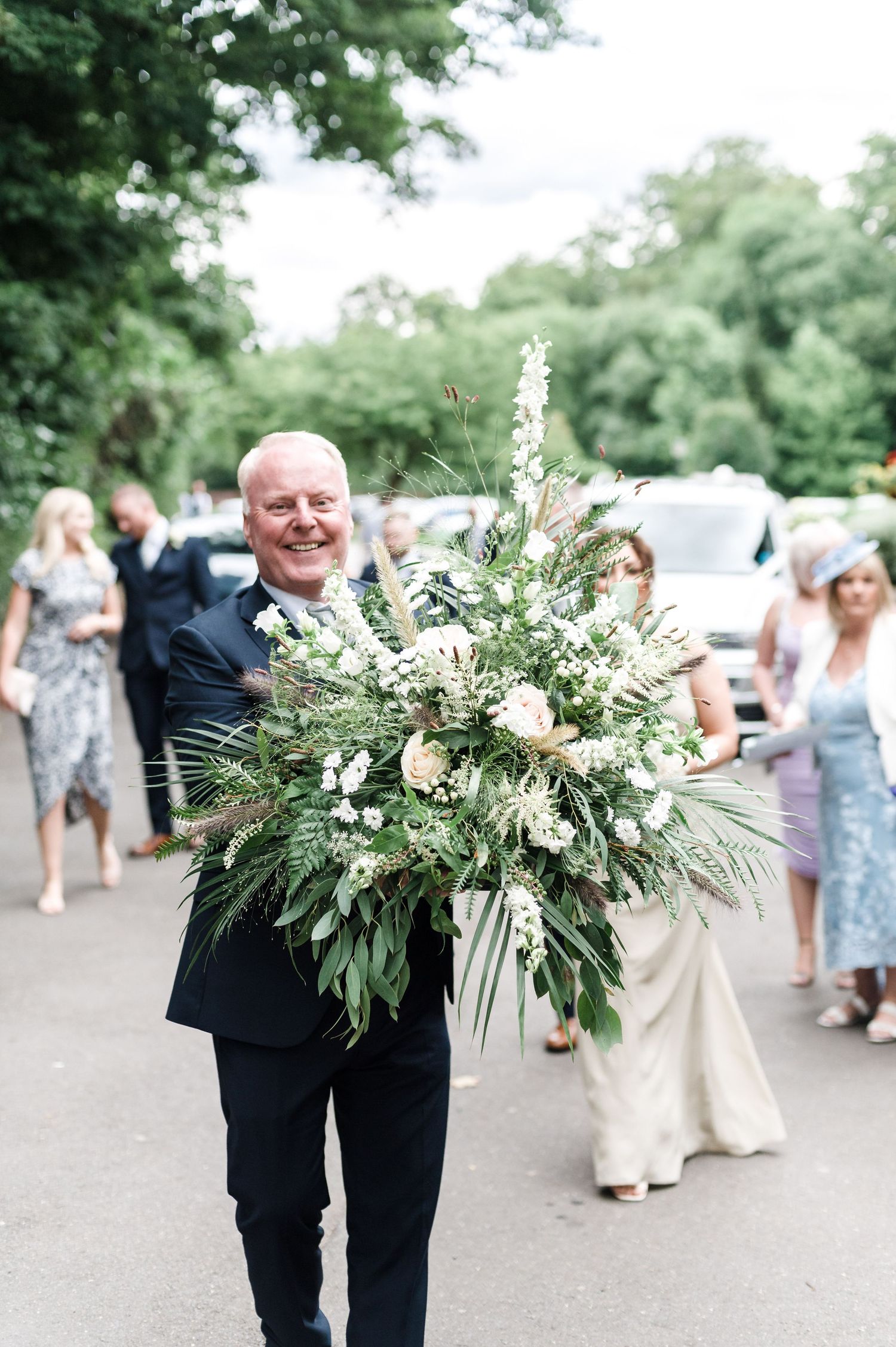 A suited individual carrying a large, lush green and white floral arrangement at an outdoor event.