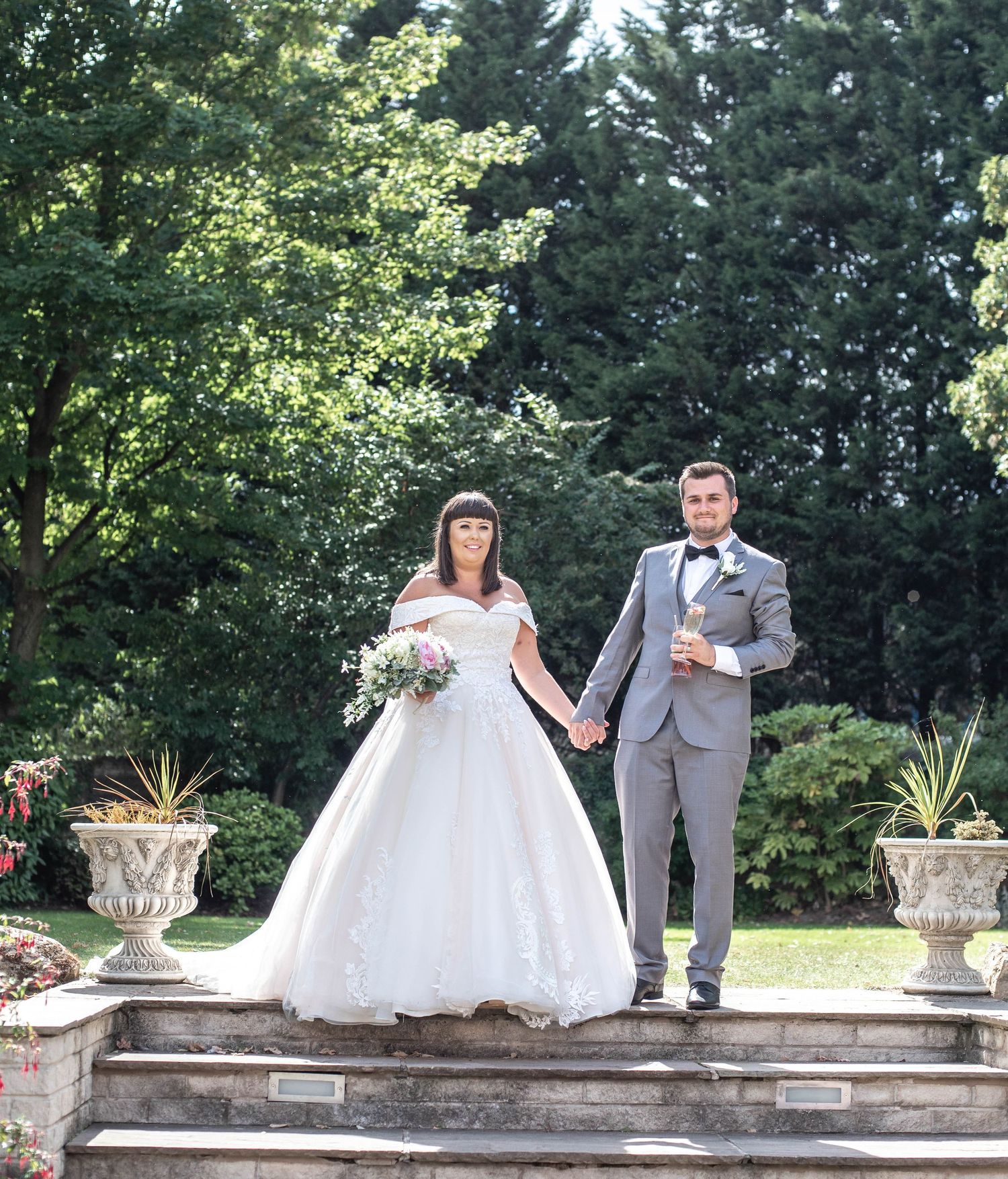 Newlyweds pose on stone steps in a garden with decorative planters.