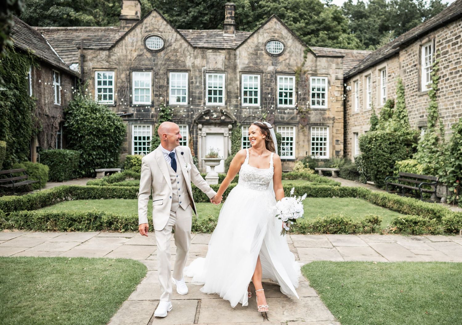 Couple walking hand in hand on garden path in front of grand stone manor house.