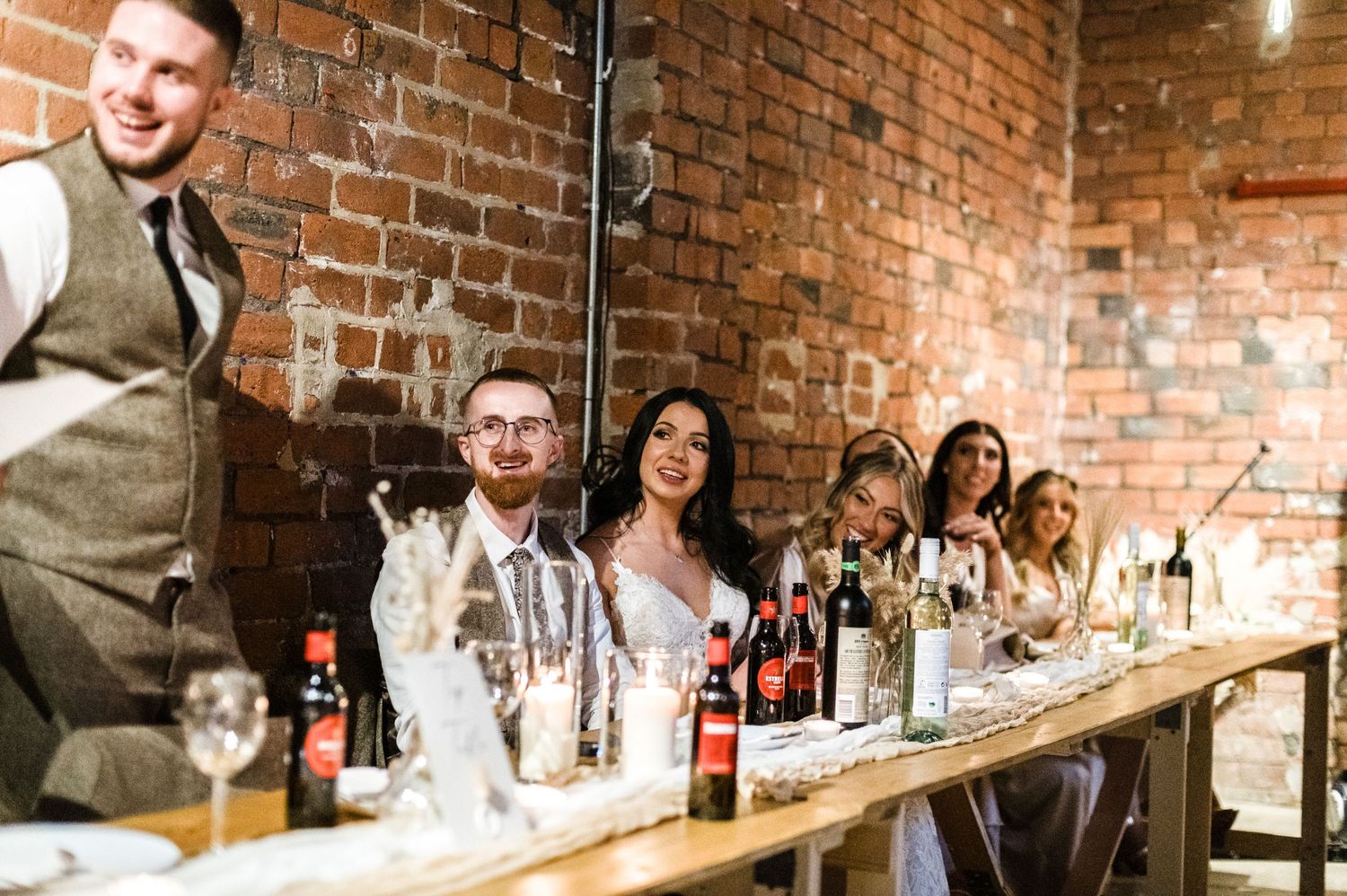 Wedding reception guests laughing and celebrating at a long dining table with brick backdrop.