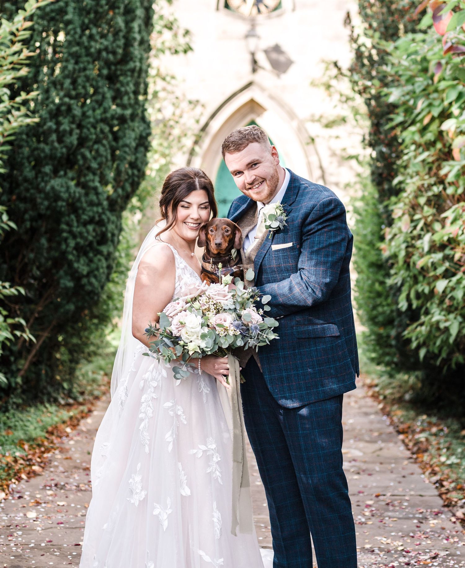 Newlyweds smile while holding their small dog in front of a gothic church entrance.