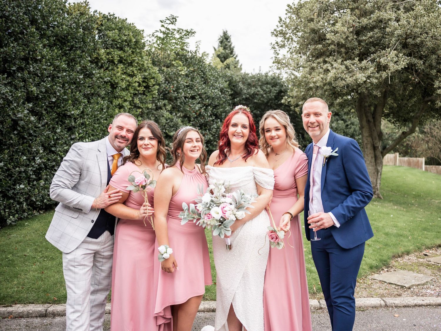 Wedding party in coordinated pink dresses and suits poses together in an outdoor garden setting.