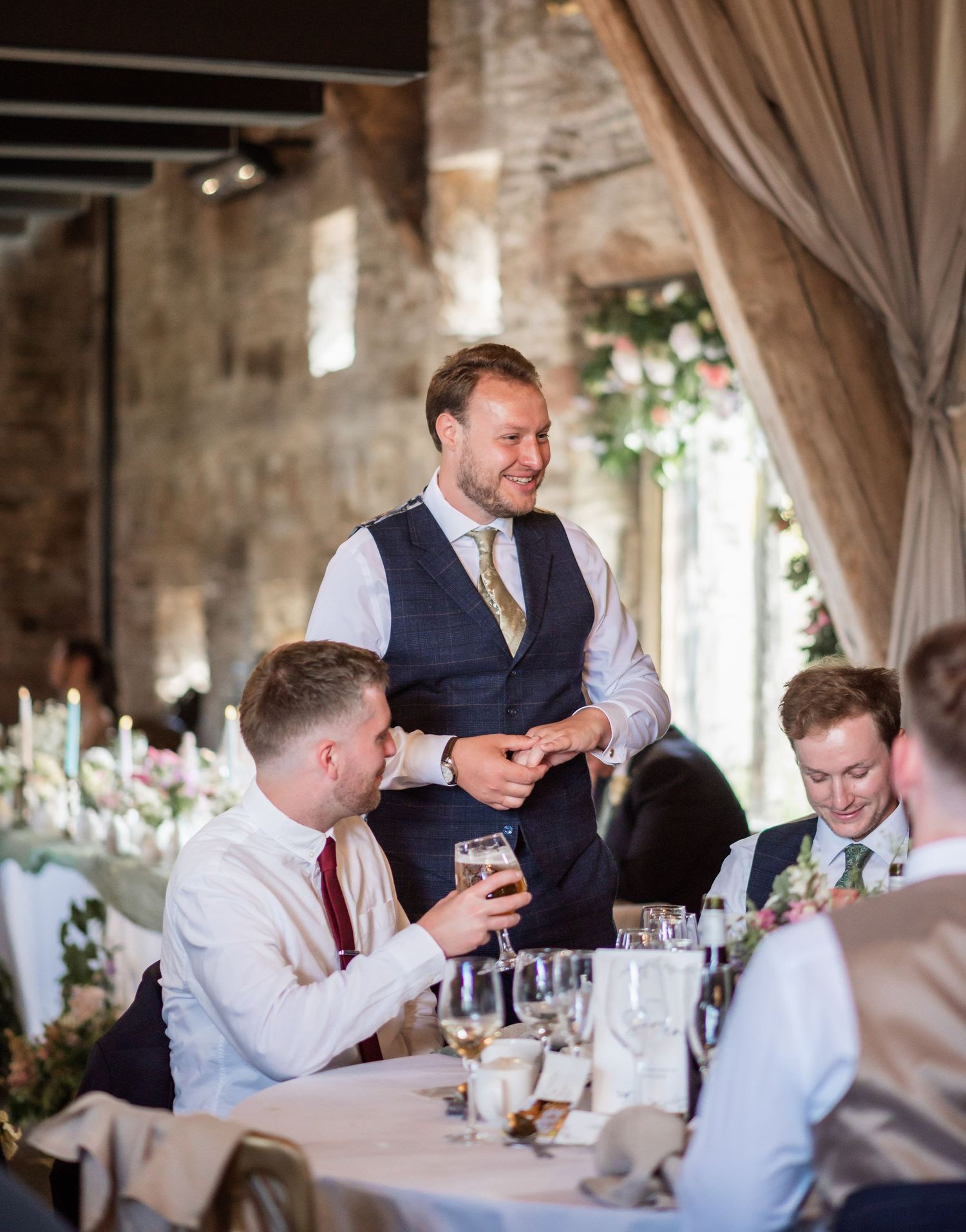 Wedding reception speech moment in rustic stone venue with guests at dining table.