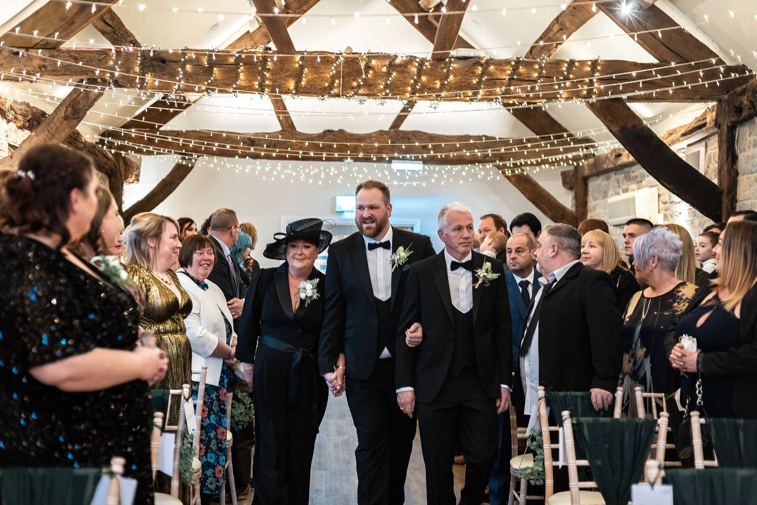 Wedding ceremony in rustic barn venue with exposed wooden beams and string lights.