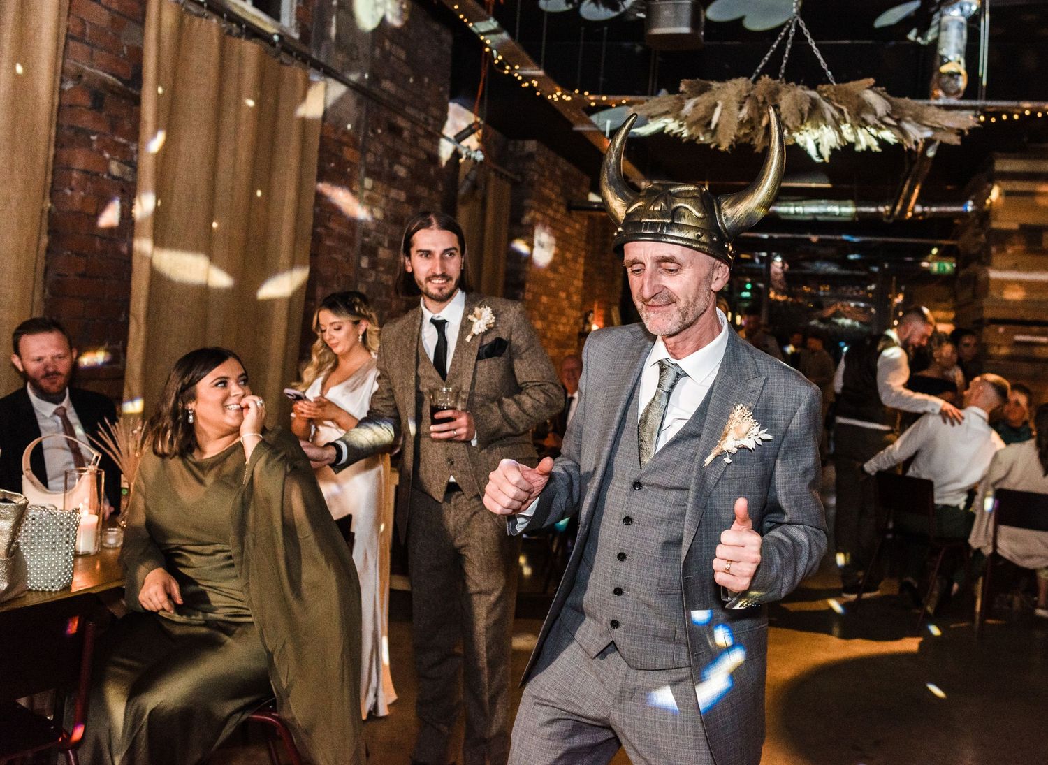 Wedding guests dancing and celebrating in an industrial venue with string lights and exposed brick walls.