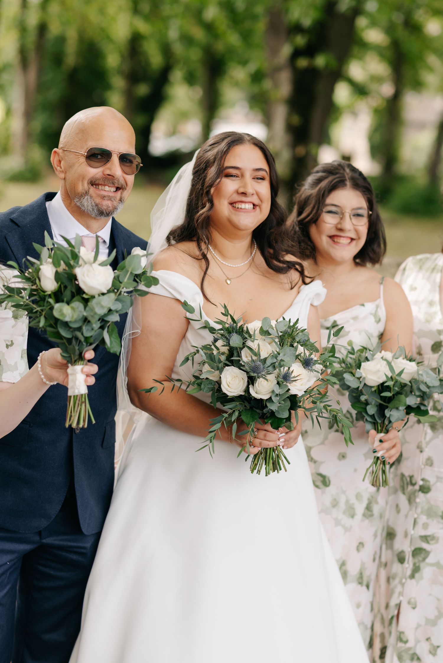 Wedding party members hold elegant white floral bouquets with greenery while posing outdoors in formal attire.