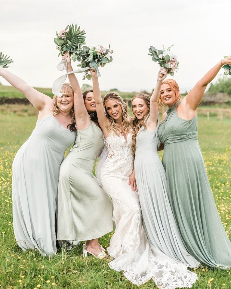 Bridesmaids in sage green dresses and bride in white celebrate joyfully in a sunny meadow.