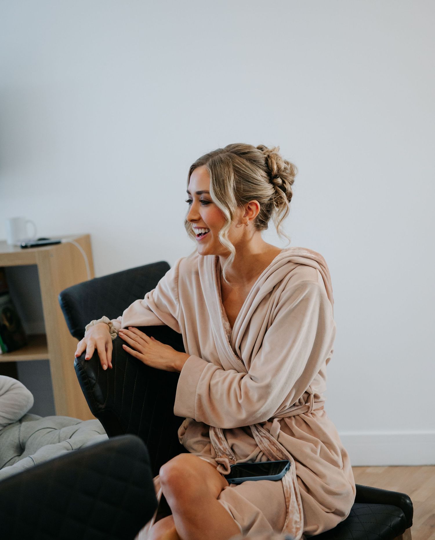 A person in a beige sweater laughs joyfully while sitting on a couch during a casual indoor photoshoot.