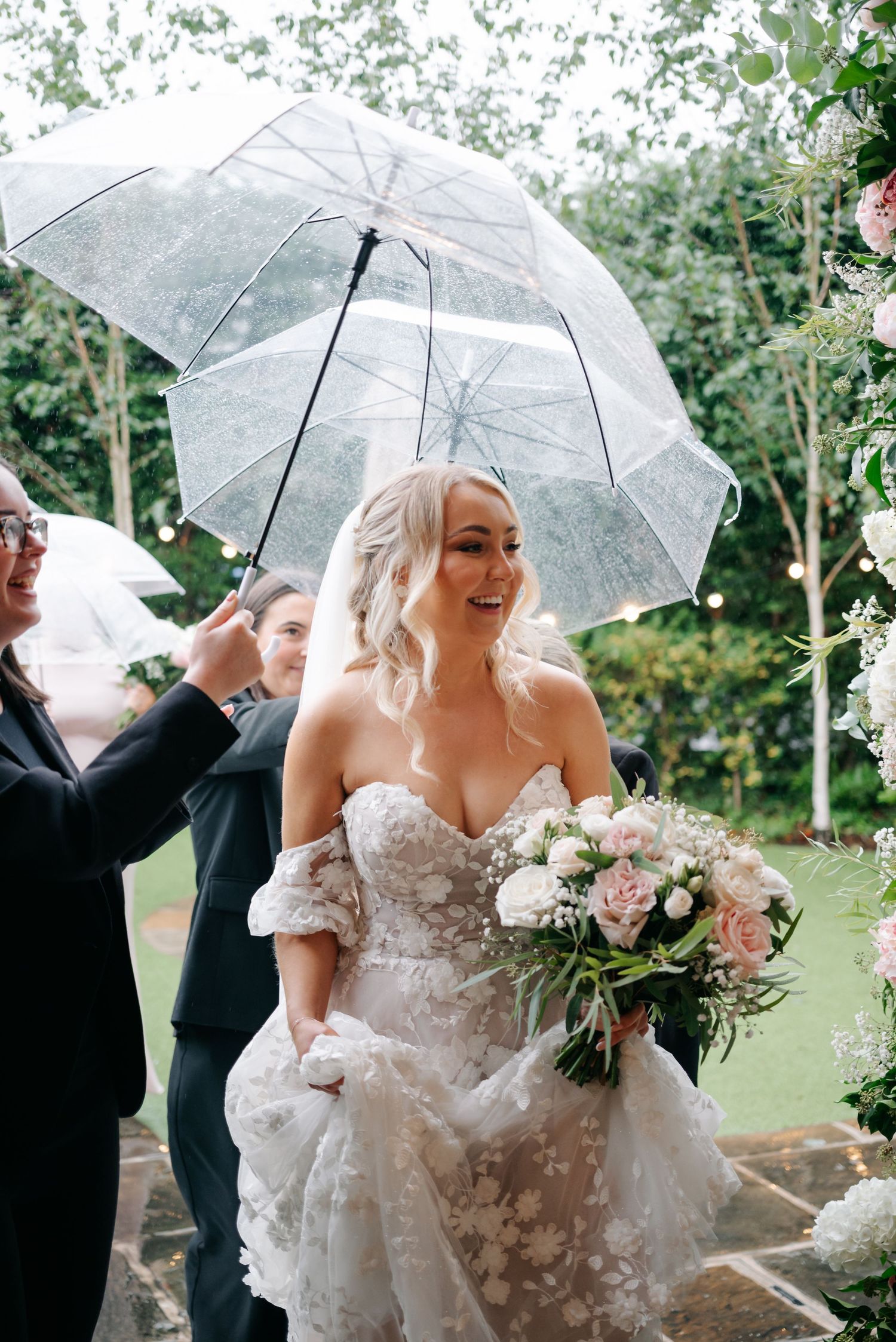 Wedding party members holding clear umbrellas while walking outdoors on a rainy day.