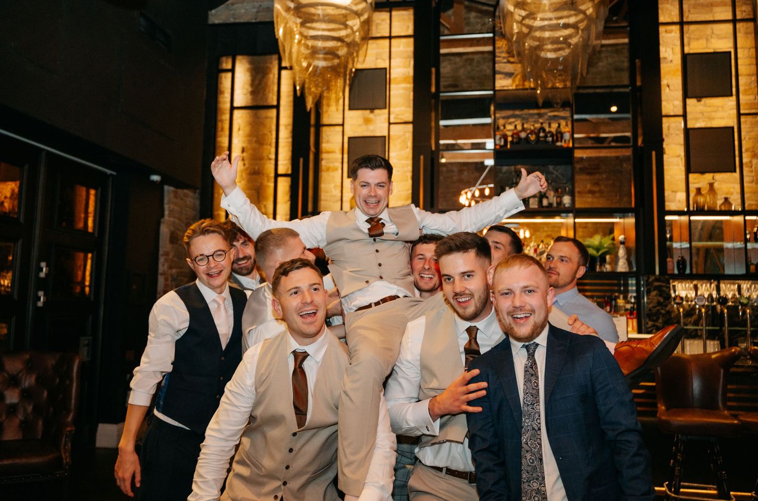 Groomsmen and guests celebrating at an indoor wedding reception with warm lighting.