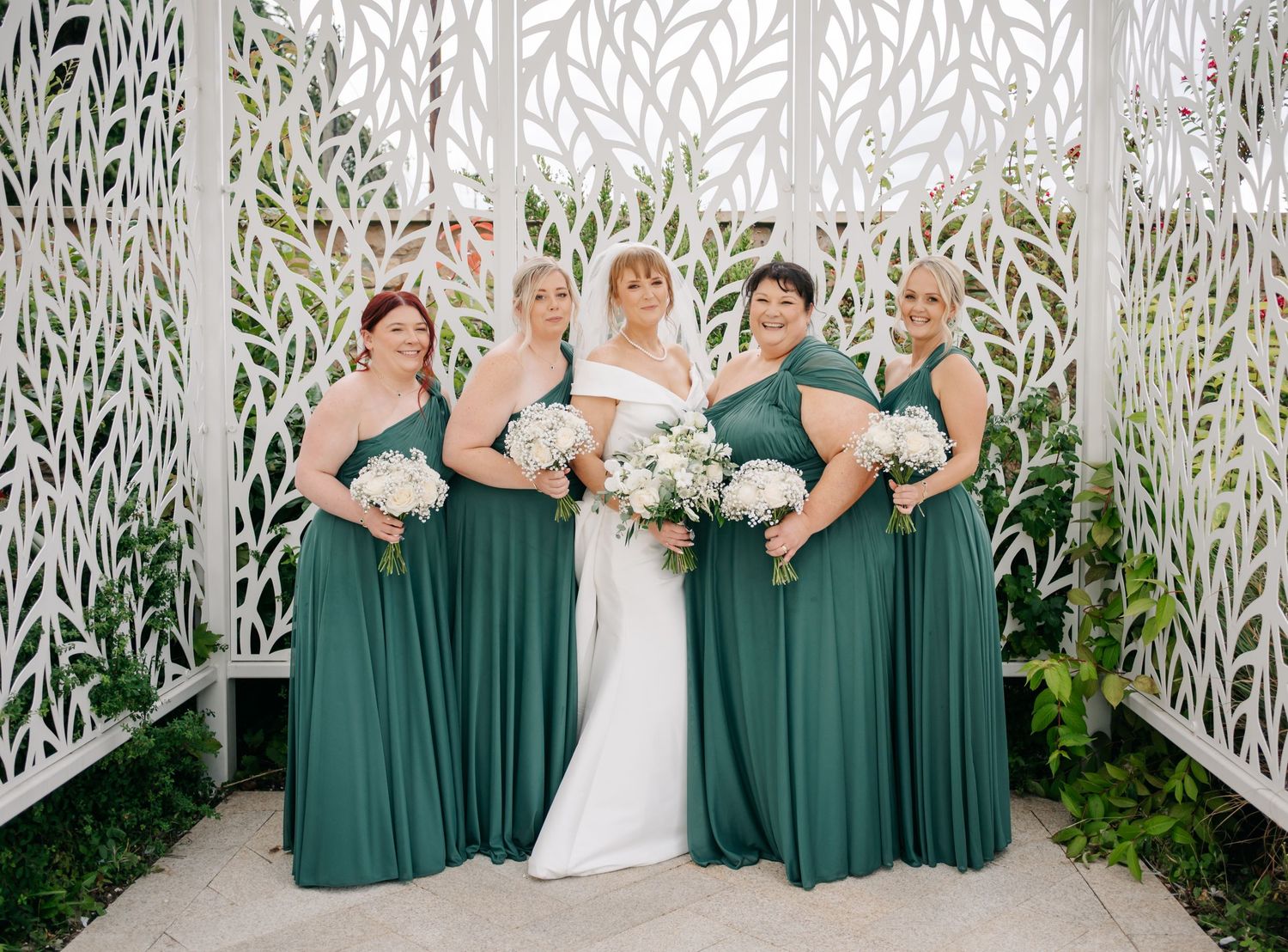 Bridesmaids in emerald green dresses stand with bride in white gown against ornate white lattice backdrop.