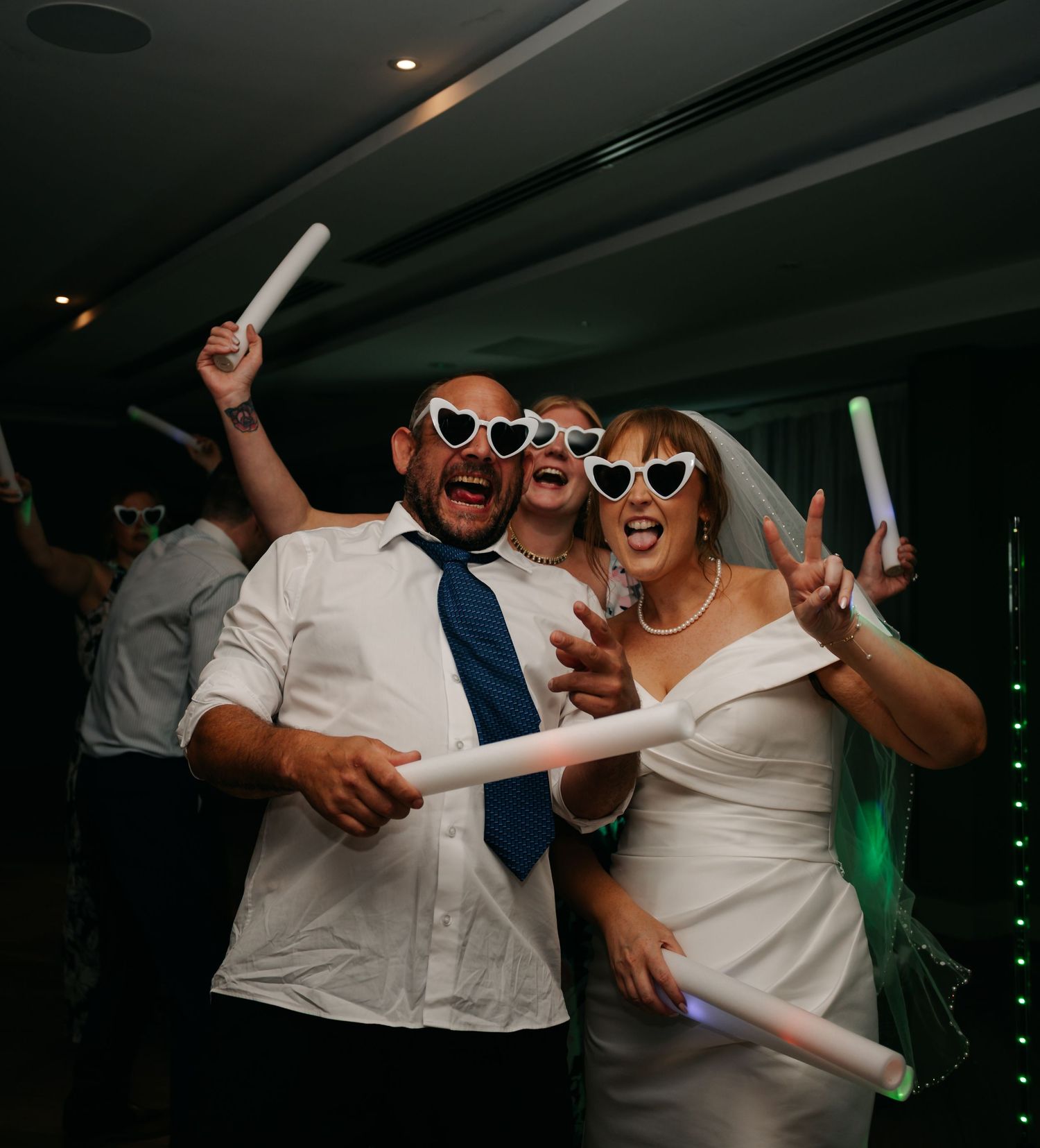 Wedding party dancing and celebrating with glow sticks in a dimly lit reception.
