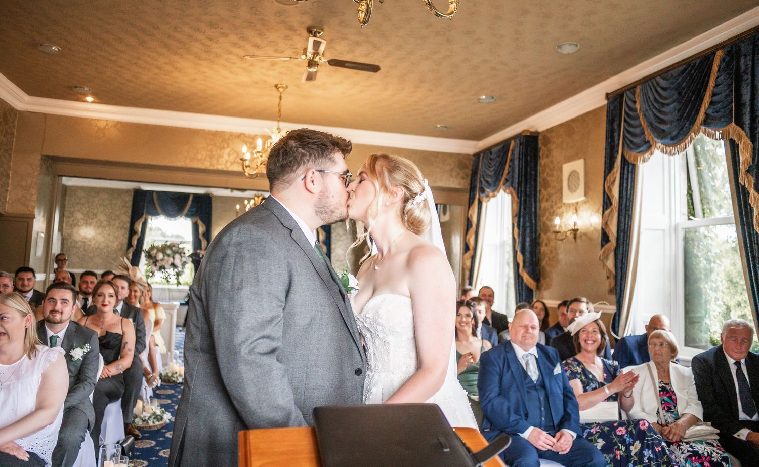 Sweet ceremony moment captured in an elegant room with draped windows and watching guests.