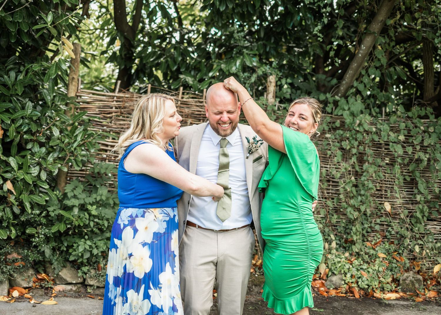 Three wedding guests in bright dresses share a joyful moment in front of a woven garden fence.