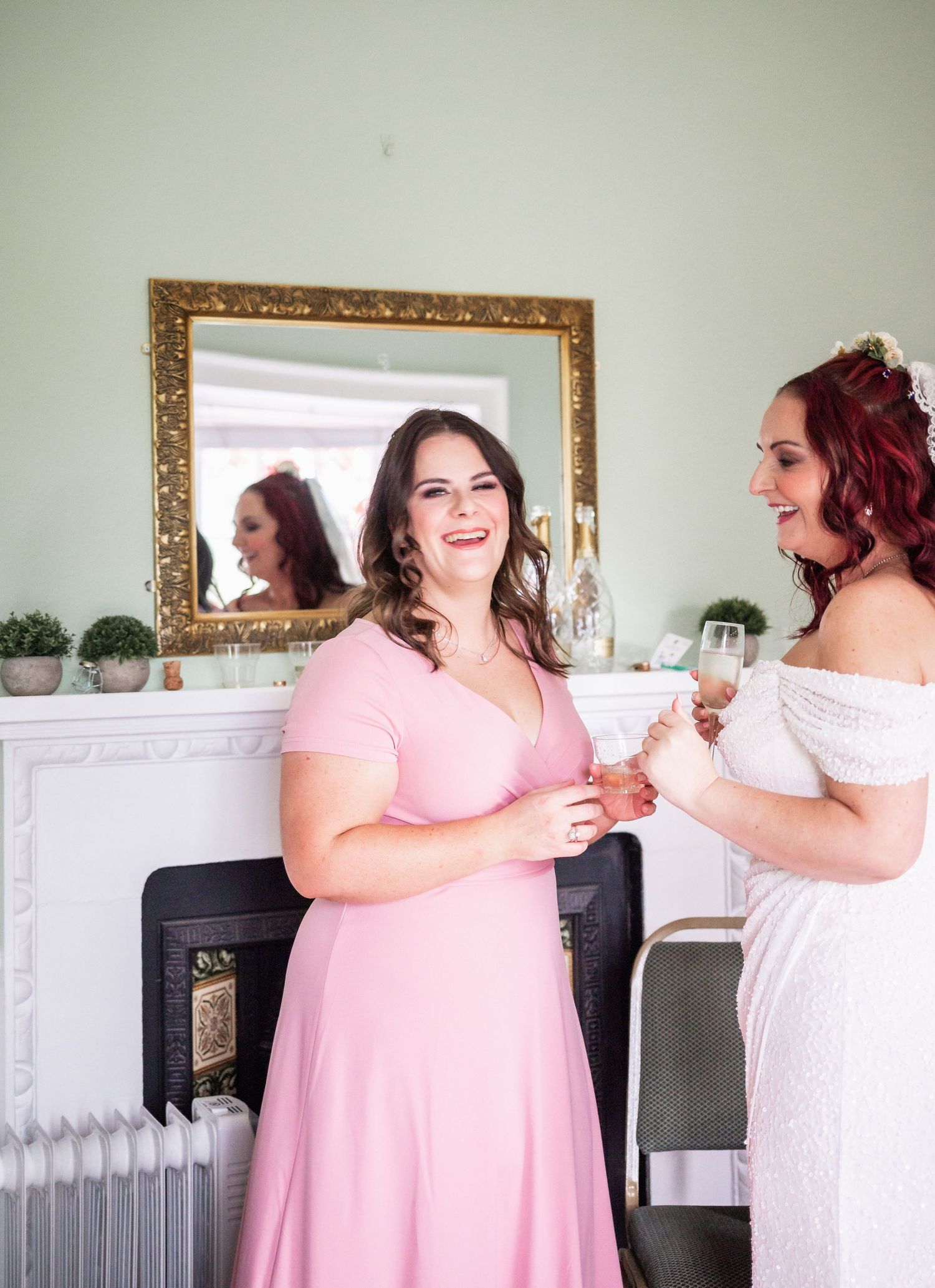 Bridesmaid in pink dress shares champagne toast with bride by ornate fireplace mirror.