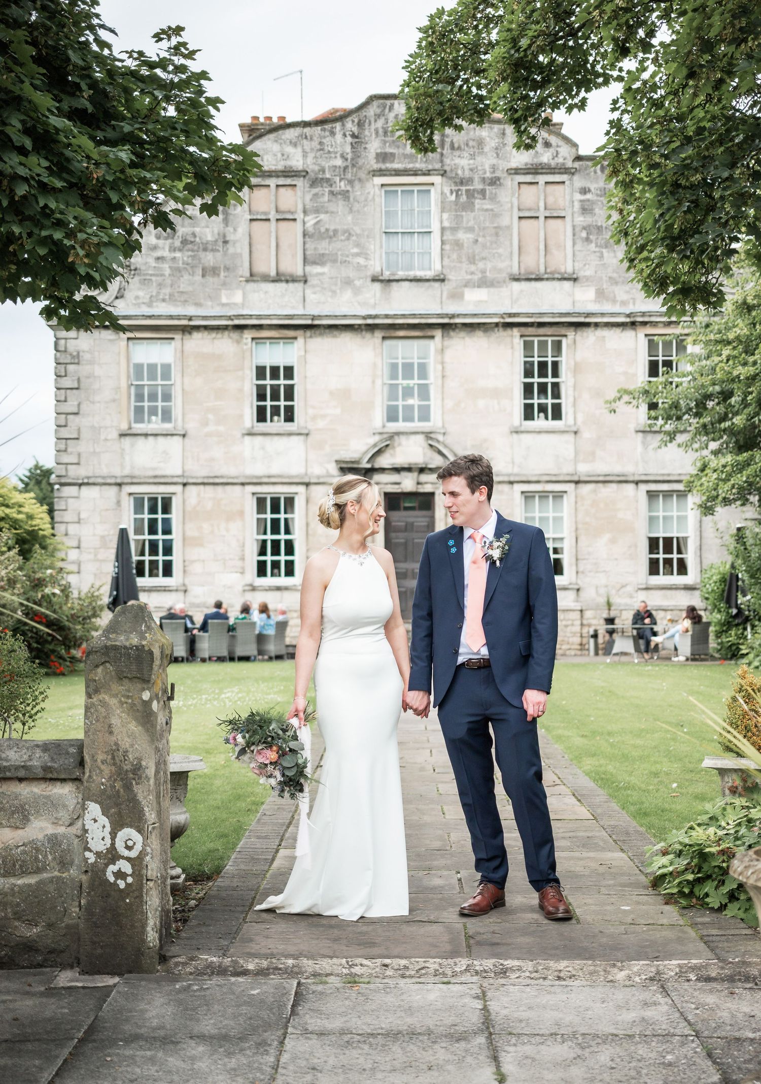 Wedding couple poses in front of elegant stone manor house on garden path.