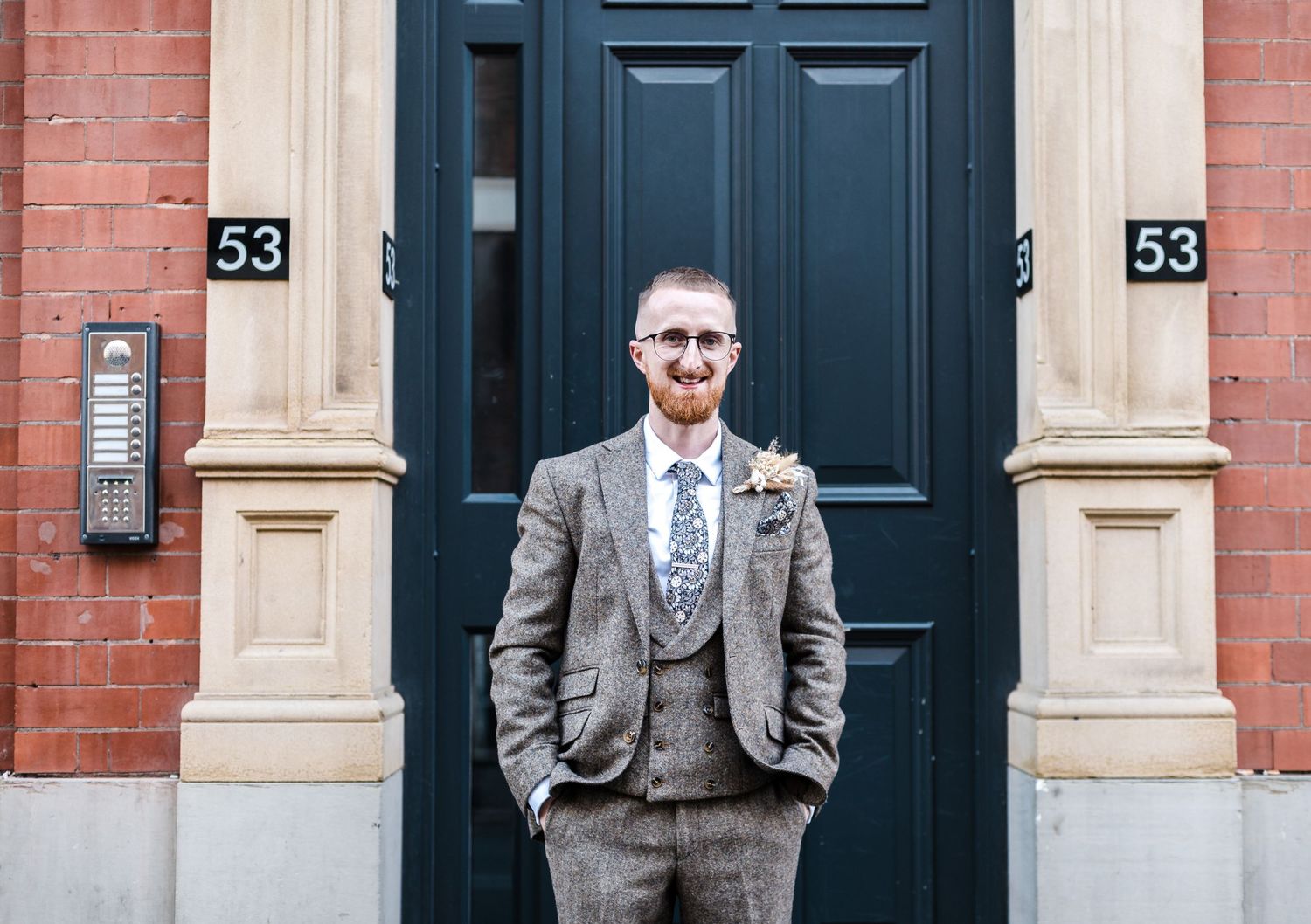 Groom in grey tweed suit stands in front of black door on brick building.