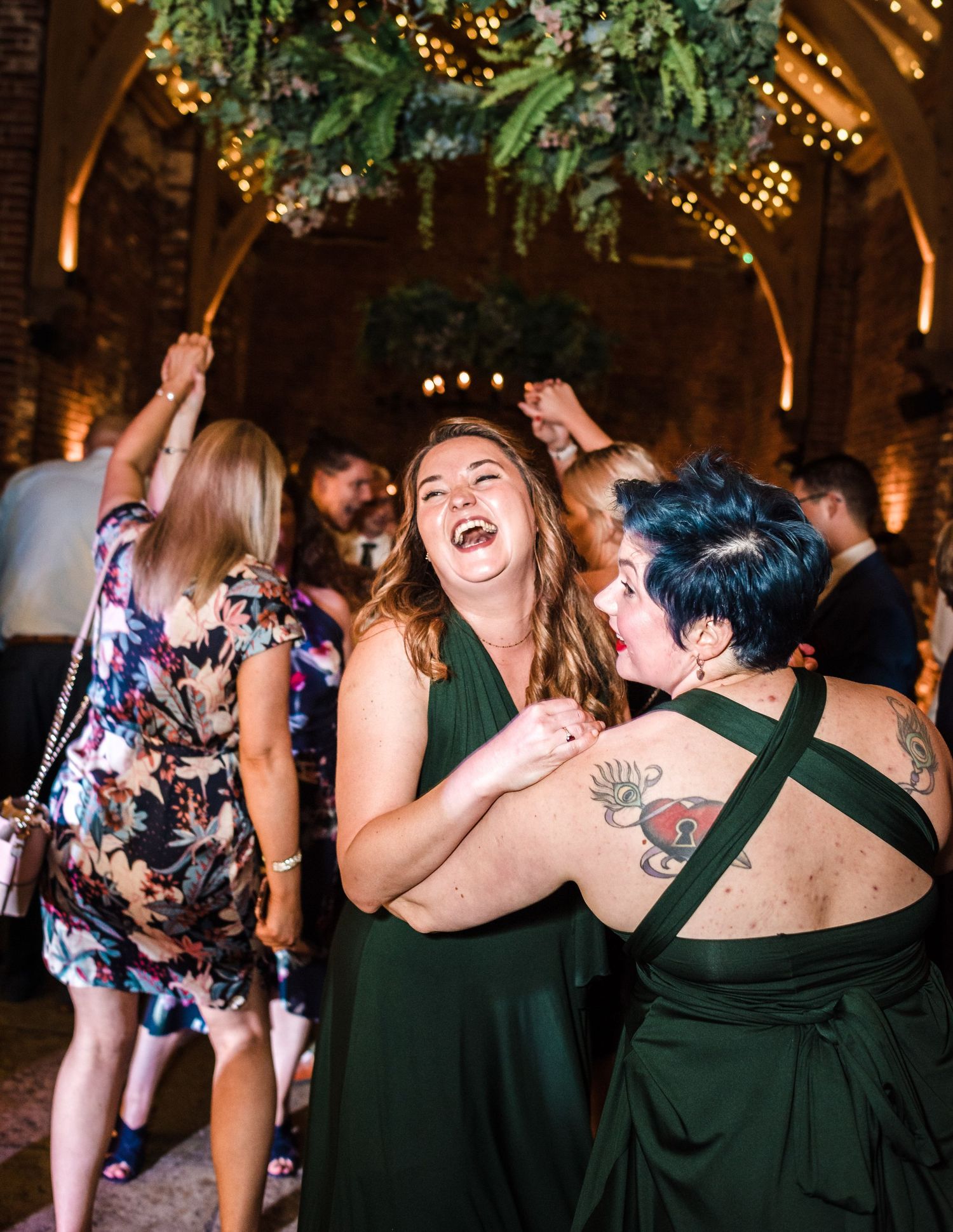 Wedding guests in colorful dresses dance and laugh joyfully at a reception.