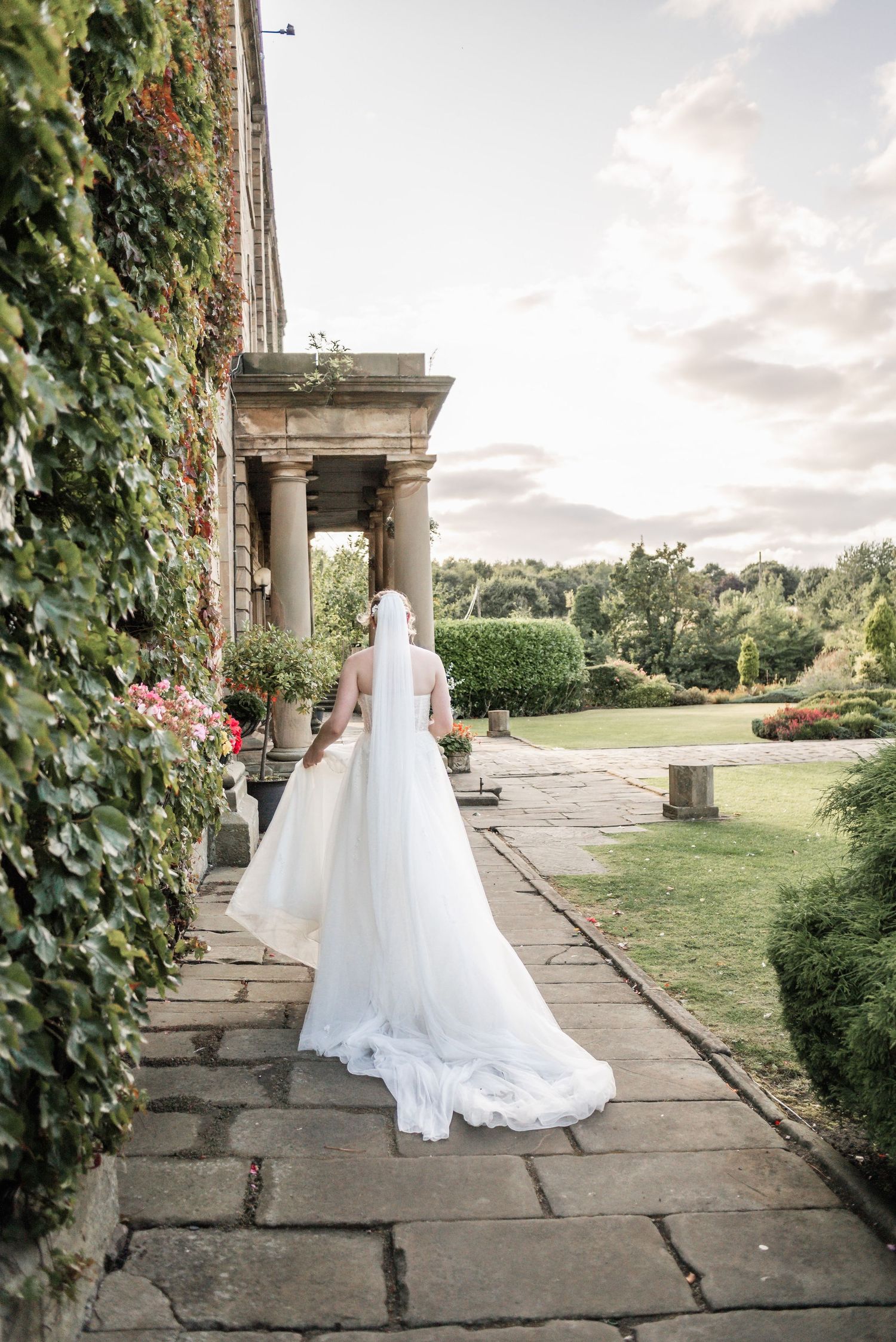 Wedding dress train flows along stone pathway by ivy-covered pillars during sunset.