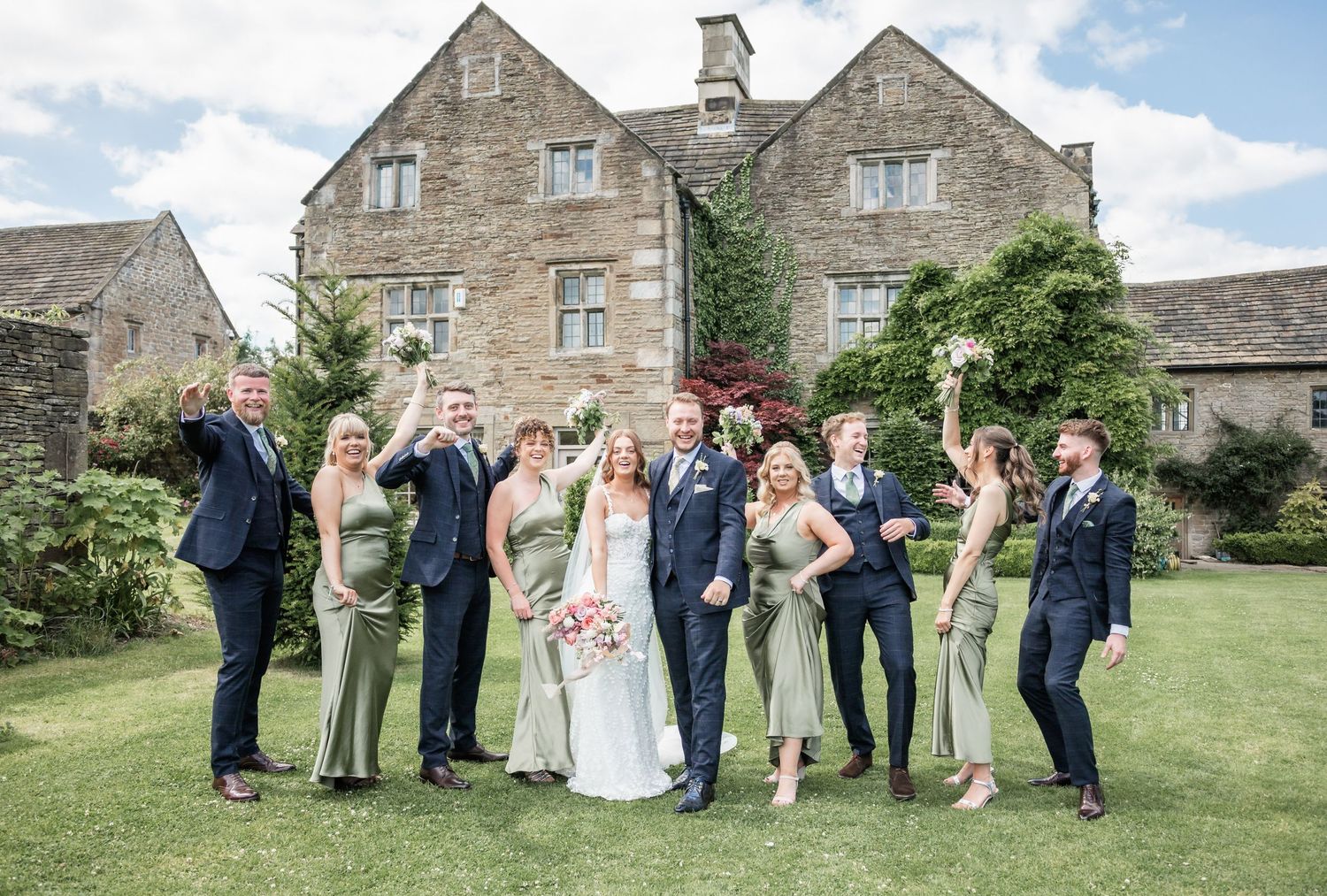 Wedding party celebrates on lawn outside stone country house.