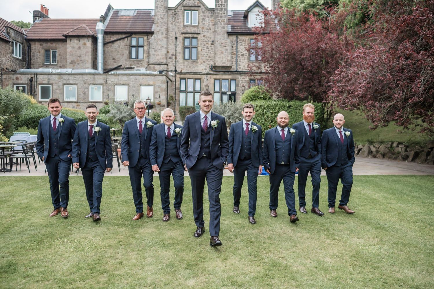 Group of groomsmen walking in navy suits on lawn in front of stone mansion.