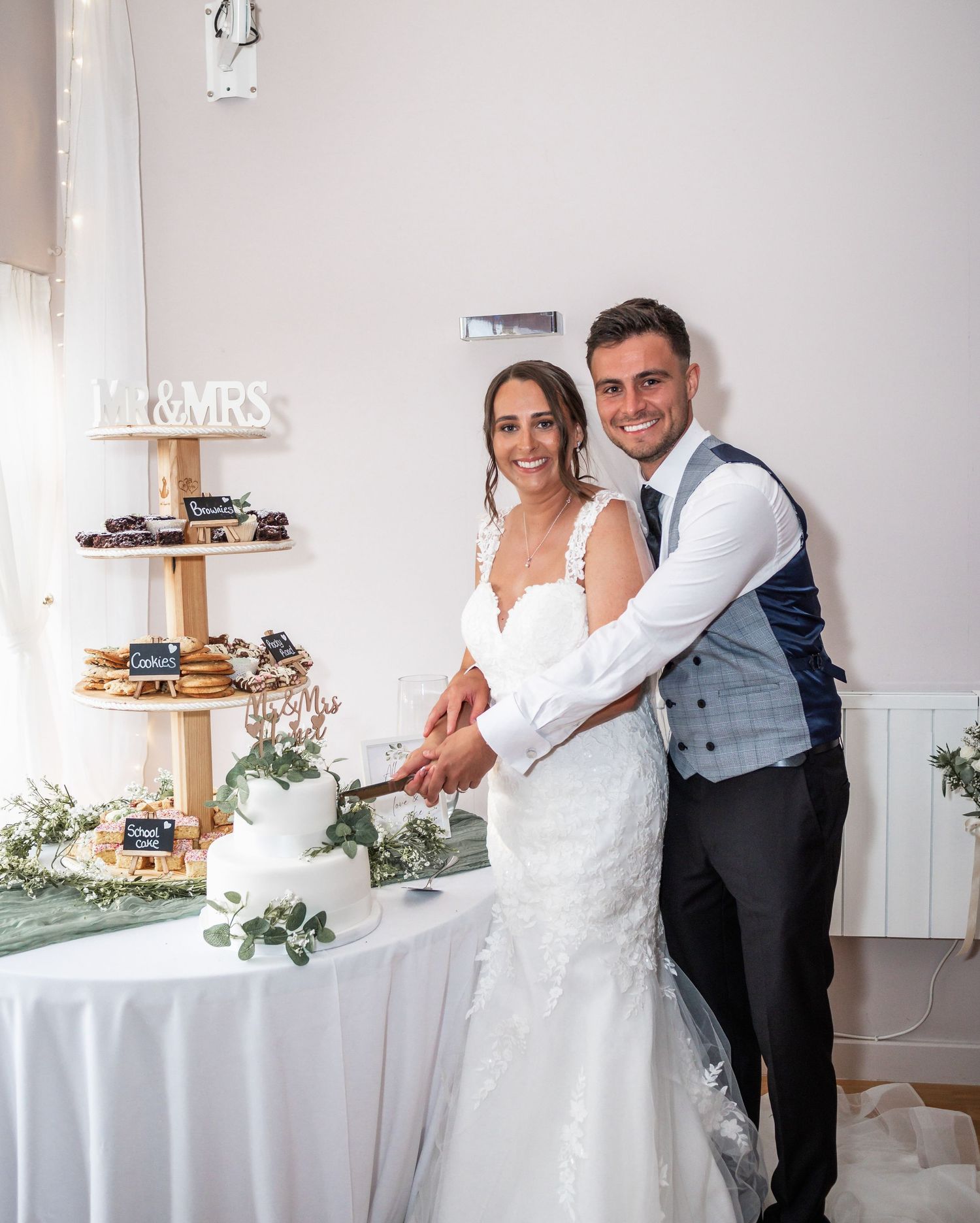 Newlyweds cutting their cake together at the dessert table decorated with greenery.