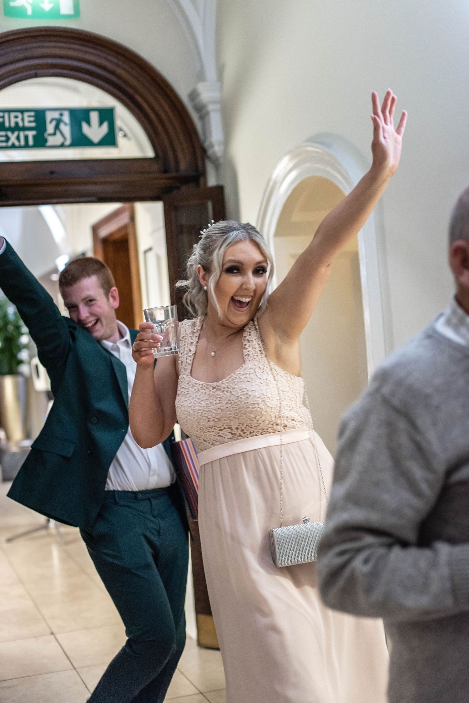 Wedding guests celebrate with raised arms in a hallway near a fire exit sign.