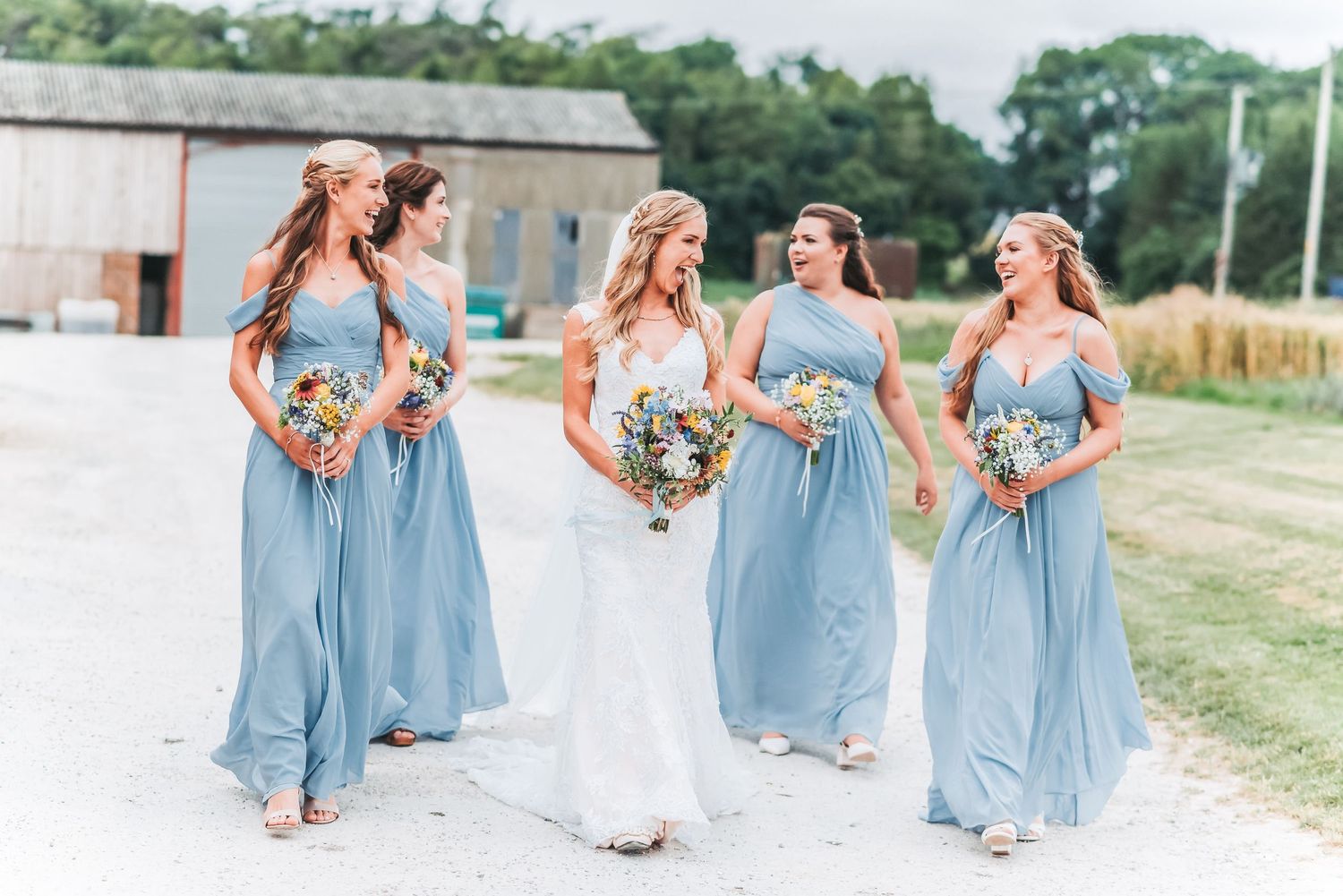 Bridal party in dusty blue dresses walking and laughing together on a rural path.
