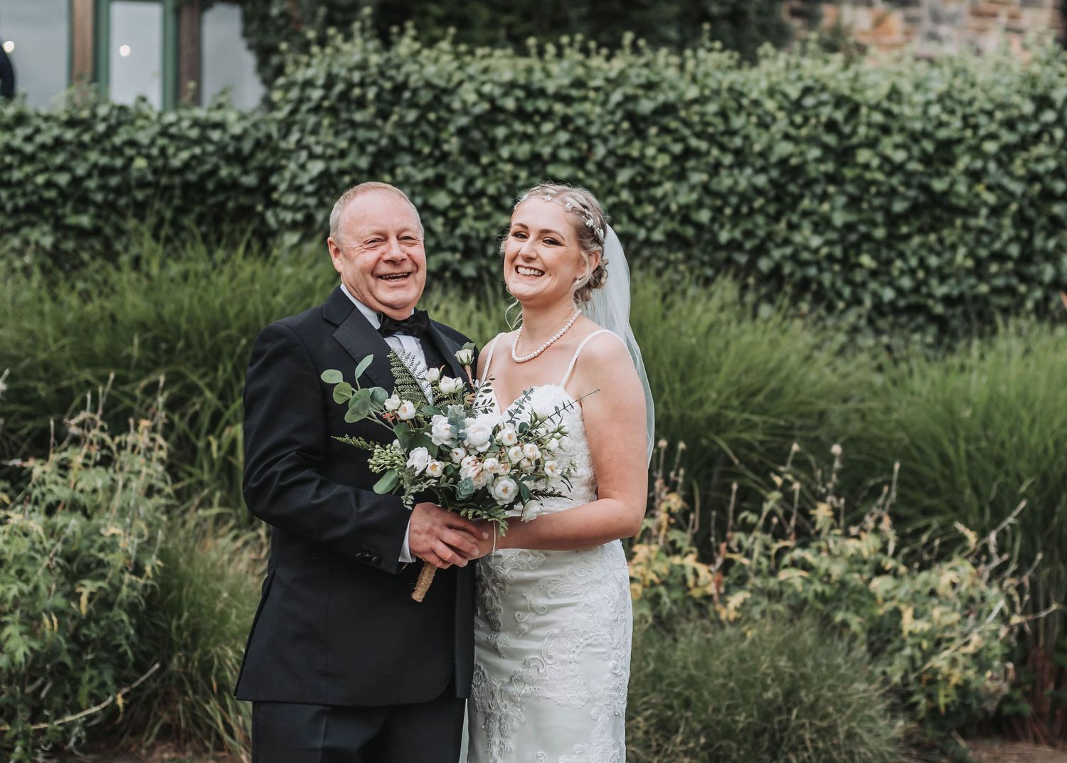 Bride poses with bouquet of white flowers in front of ivy-covered wall.