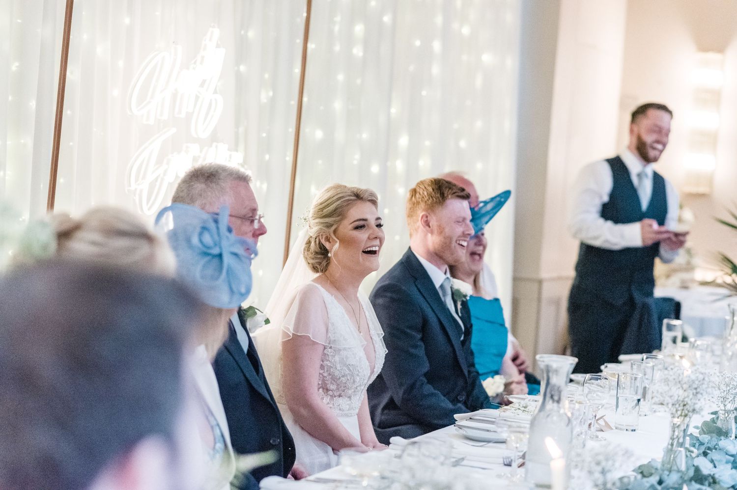 Wedding reception guests enjoying speeches and laughter at a decorated banquet table.