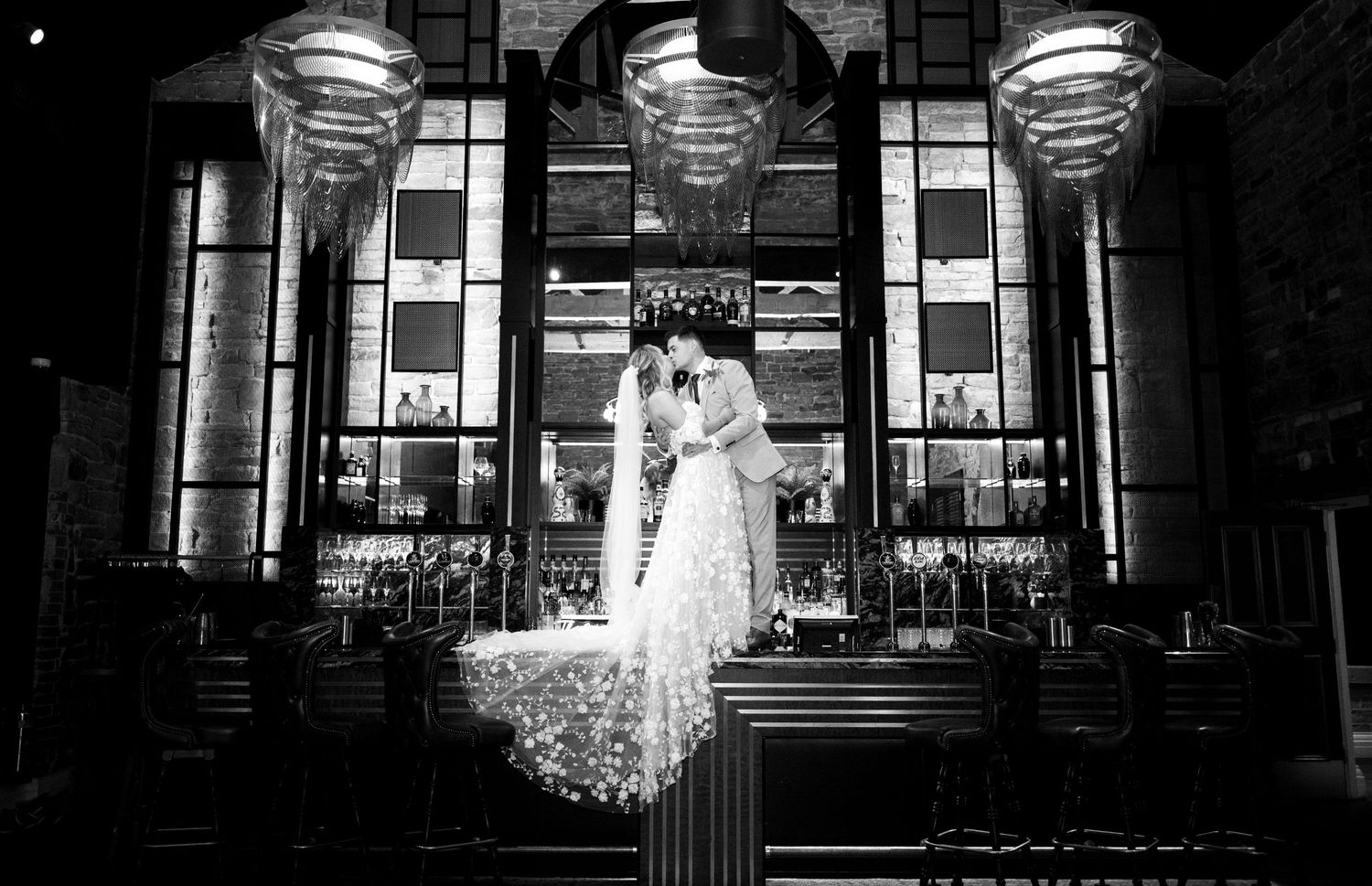 A black and white photograph of a couple in formal attire standing in front of an elegantly lit art deco building entrance.