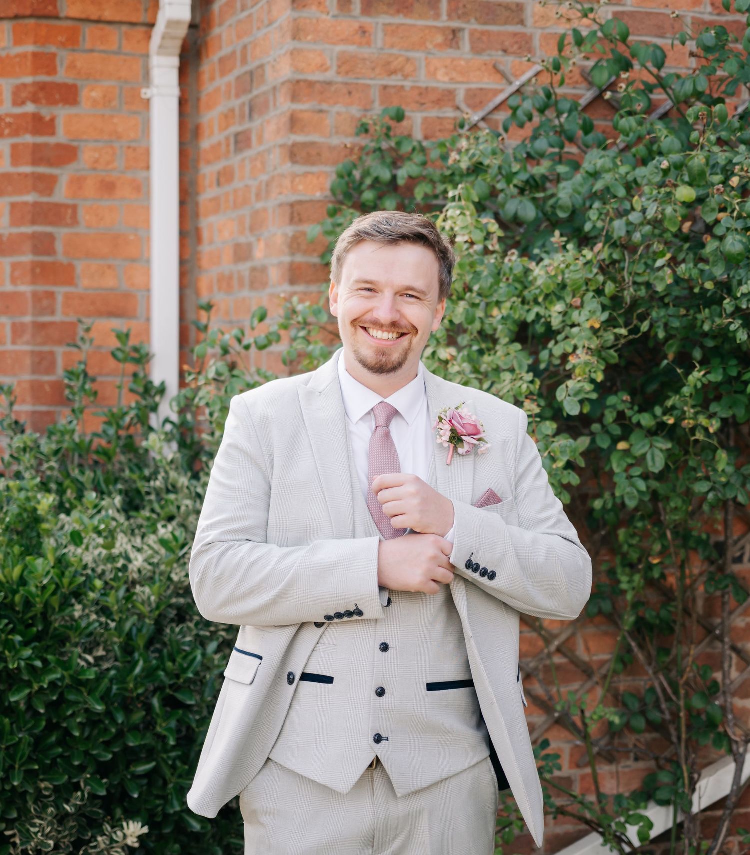 A groomsman in a light beige suit and pink tie poses outdoors against a red brick wall with greenery.