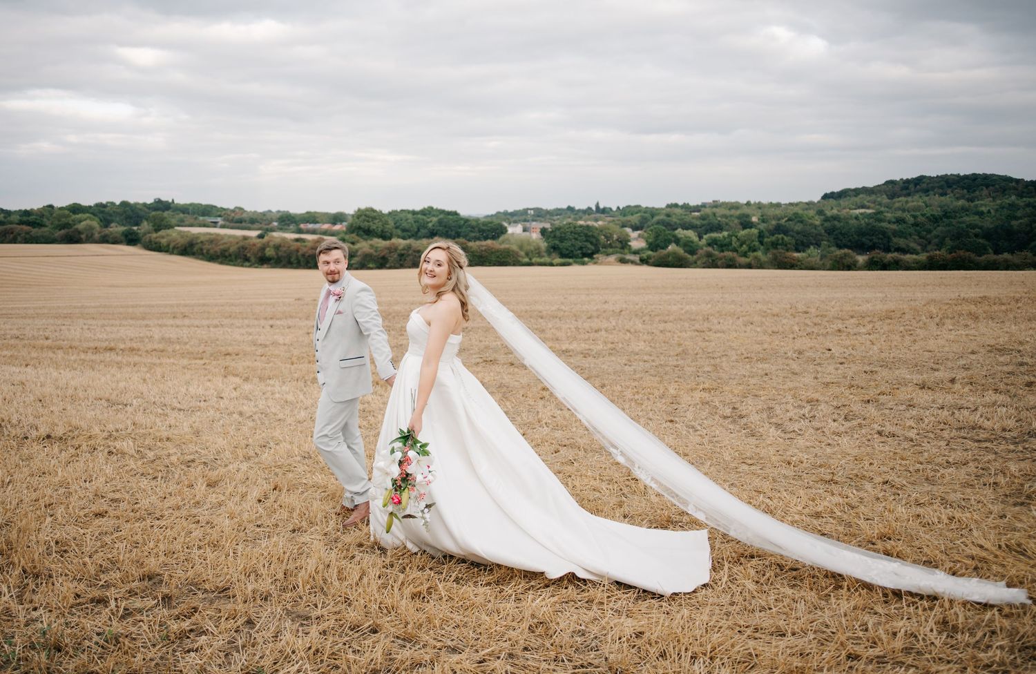 Sweeping countryside landscape with wedding couple walking through golden wheat fields.