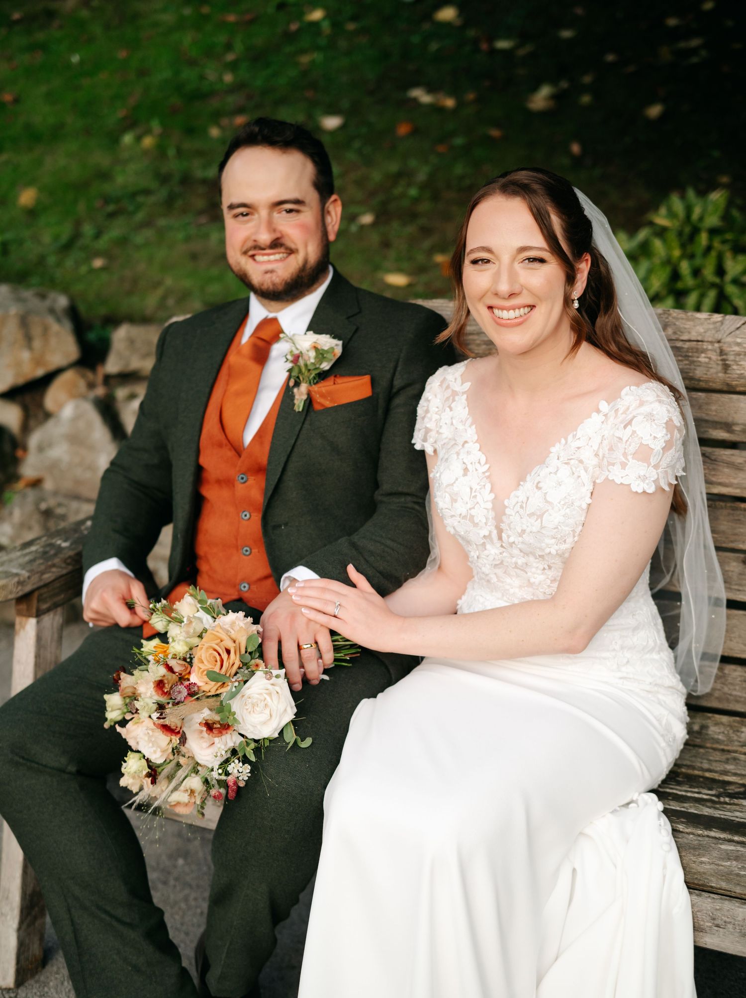 A happy bride and groom sit together on a wooden bench, holding a rustic autumn bouquet of cream and orange flowers.