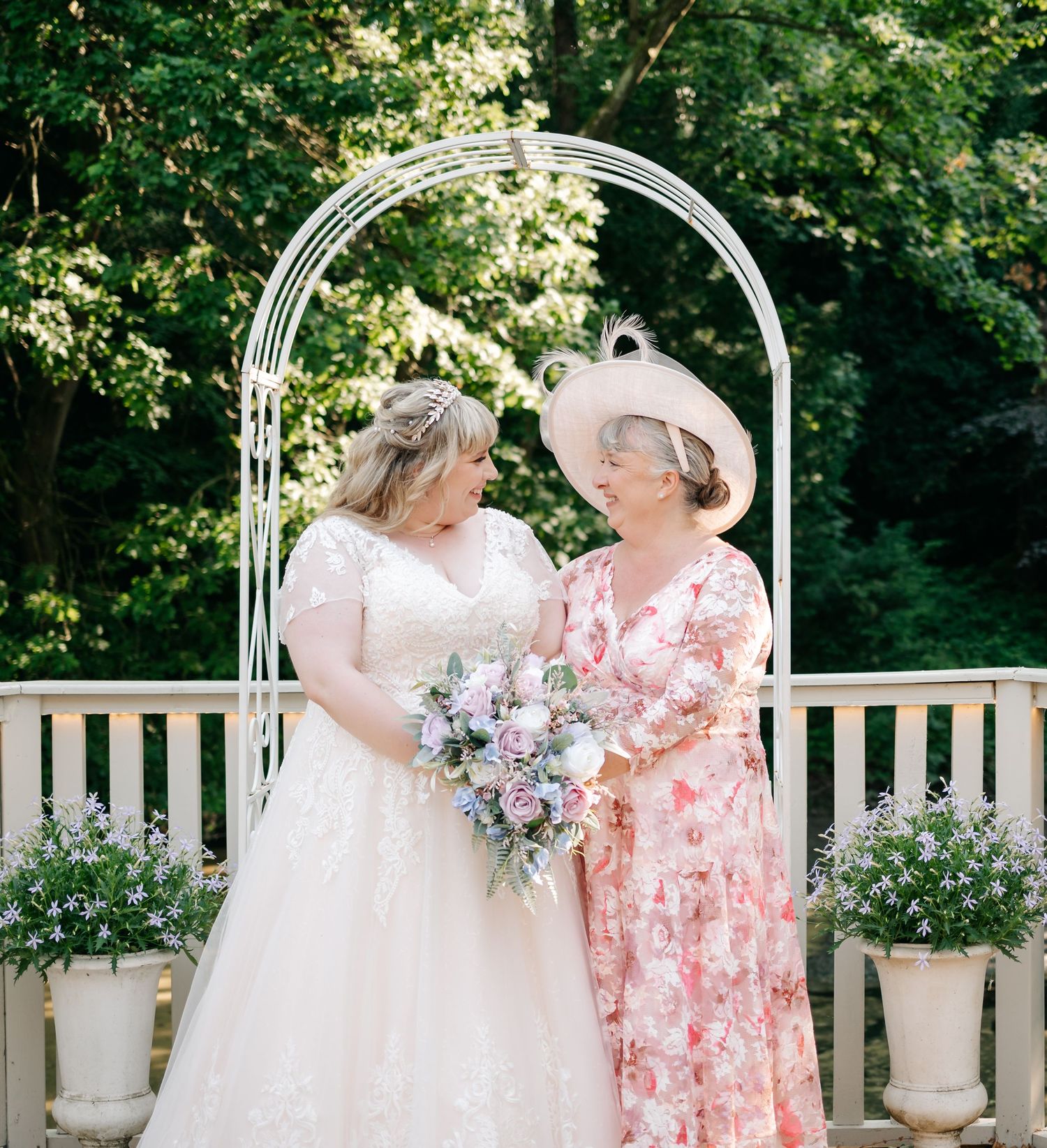 Bride in lace gown holding pastel bouquet shares a joyful moment with her mother in floral dress outdoors.