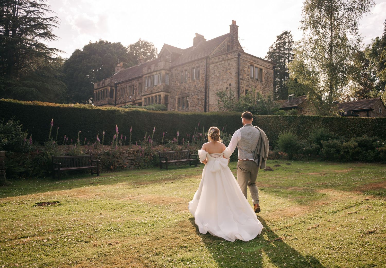 Couple walking hand in hand across sunny lawn with stone buildings in background.