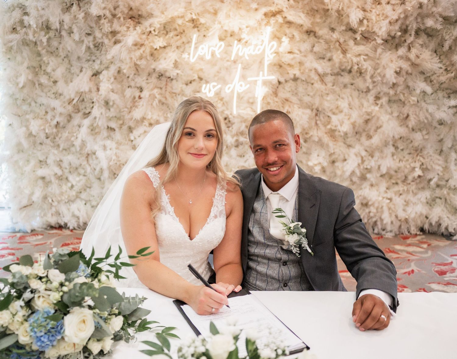Couple signs marriage documents beneath neon sign with elegant floral arrangements.