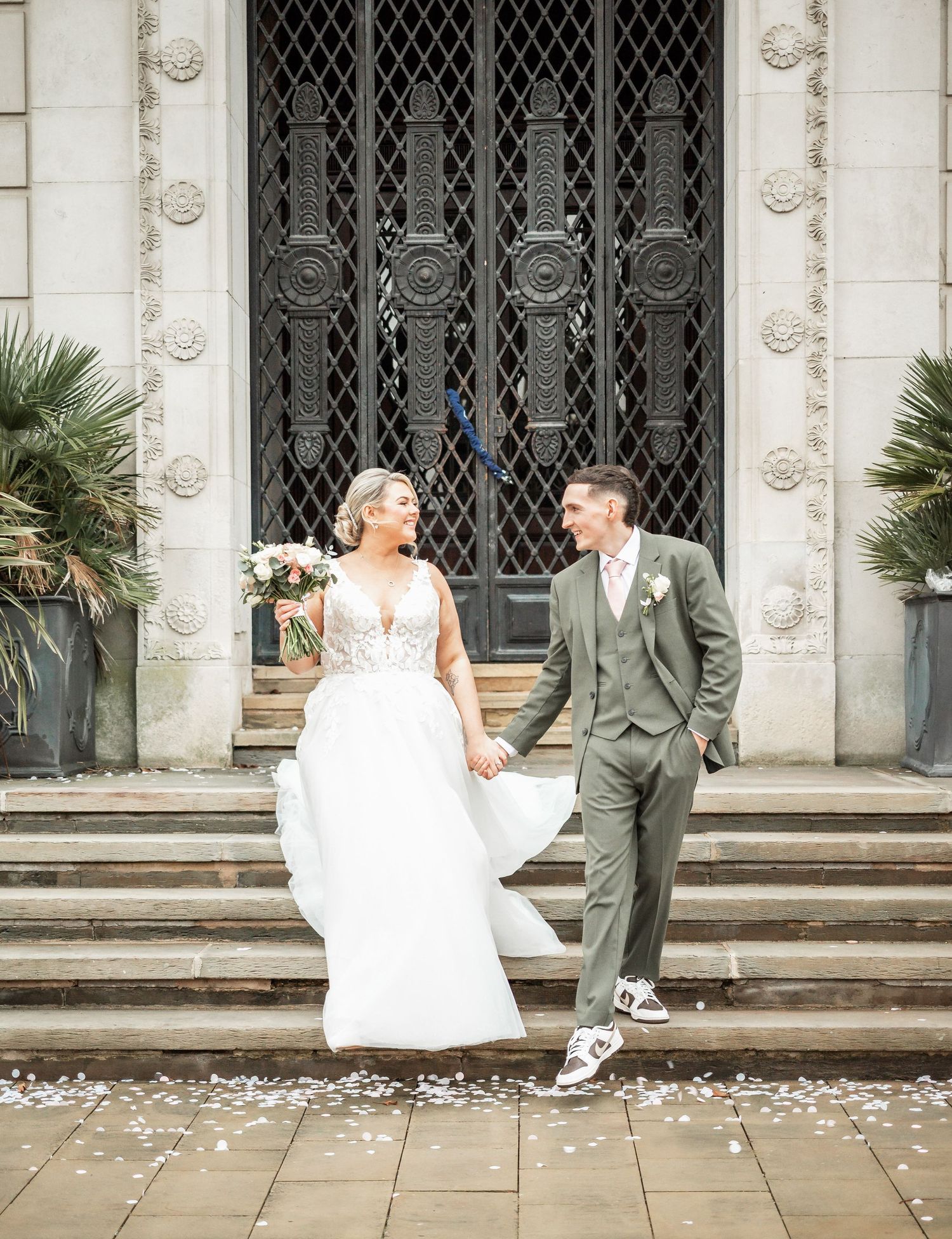 Wedding couple shares candid moment on stone steps with scattered petals and iron gate backdrop.