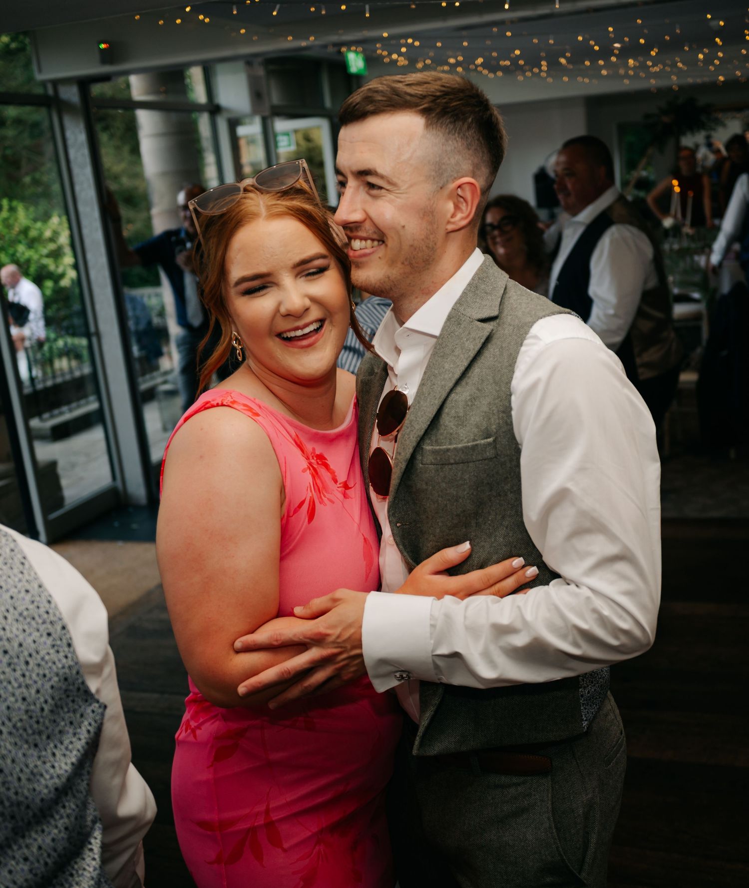 A couple shares joyful laughter during an intimate moment at an indoor venue with string lights visible in the background.