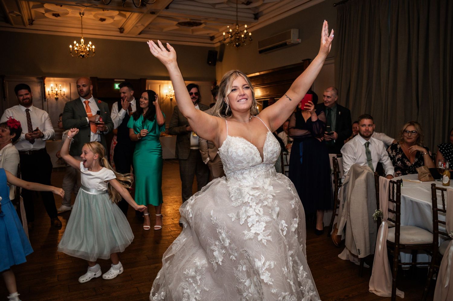 A person in a white wedding dress dances with arms raised during wedding reception celebrations.
