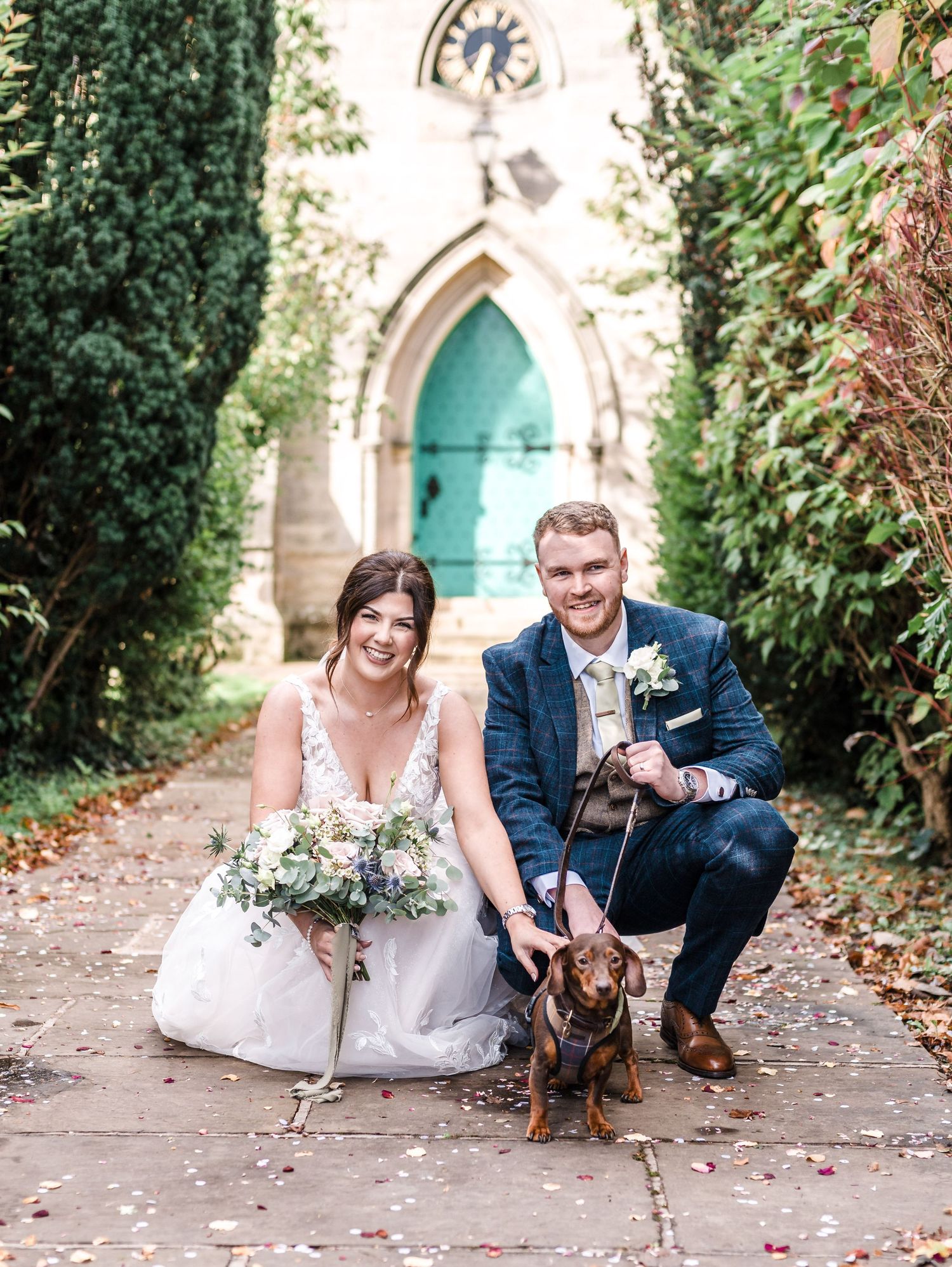 A couple poses with their small dog in front of a turquoise church door surrounded by greenery.